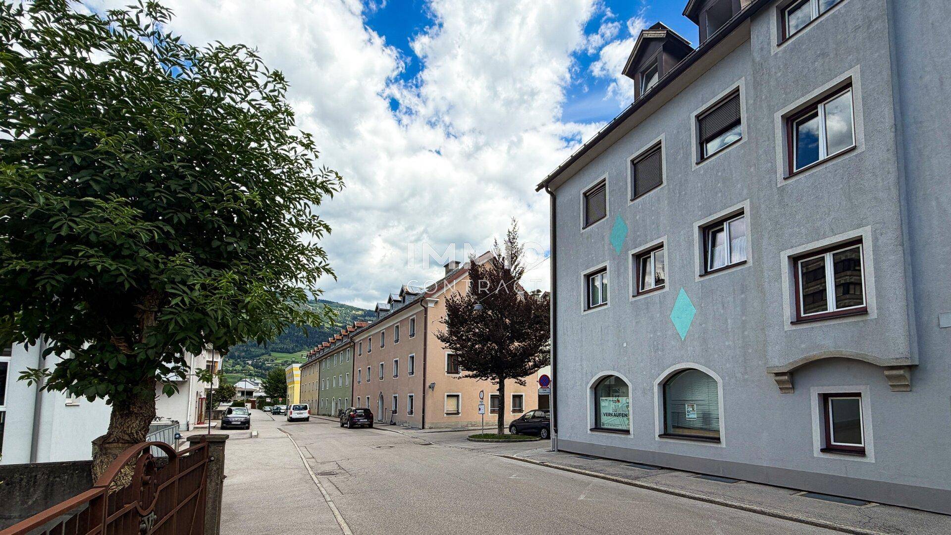 Blick auf eine Wohnstraße mit Gebäuden, Bäumen und Bergen im Hintergrund unter blauem Himmel.