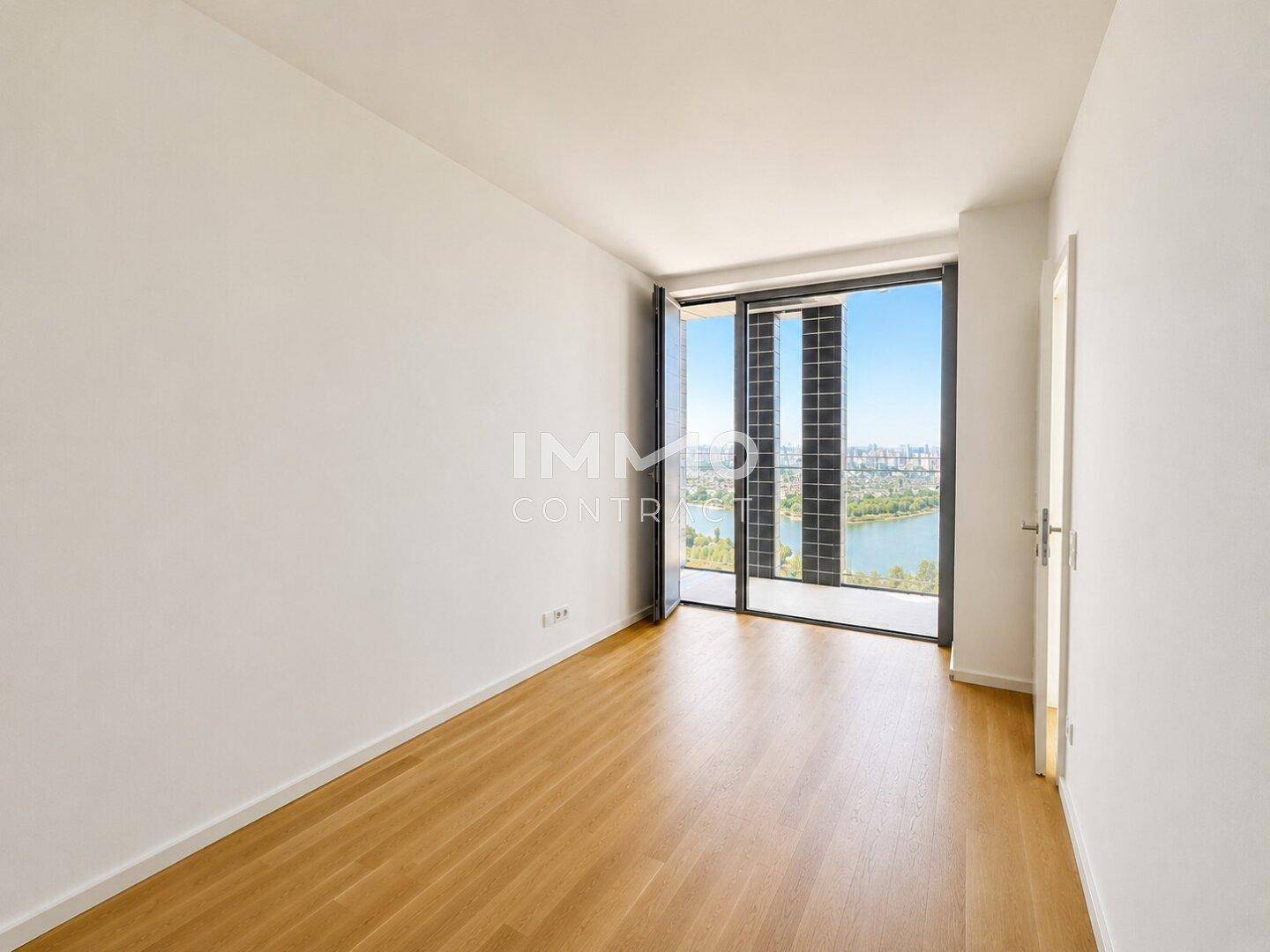 Leeres Zimmer mit Holzboden und großem Fenster, das einen weiten Blick auf die Stadt und den Fluss bietet.