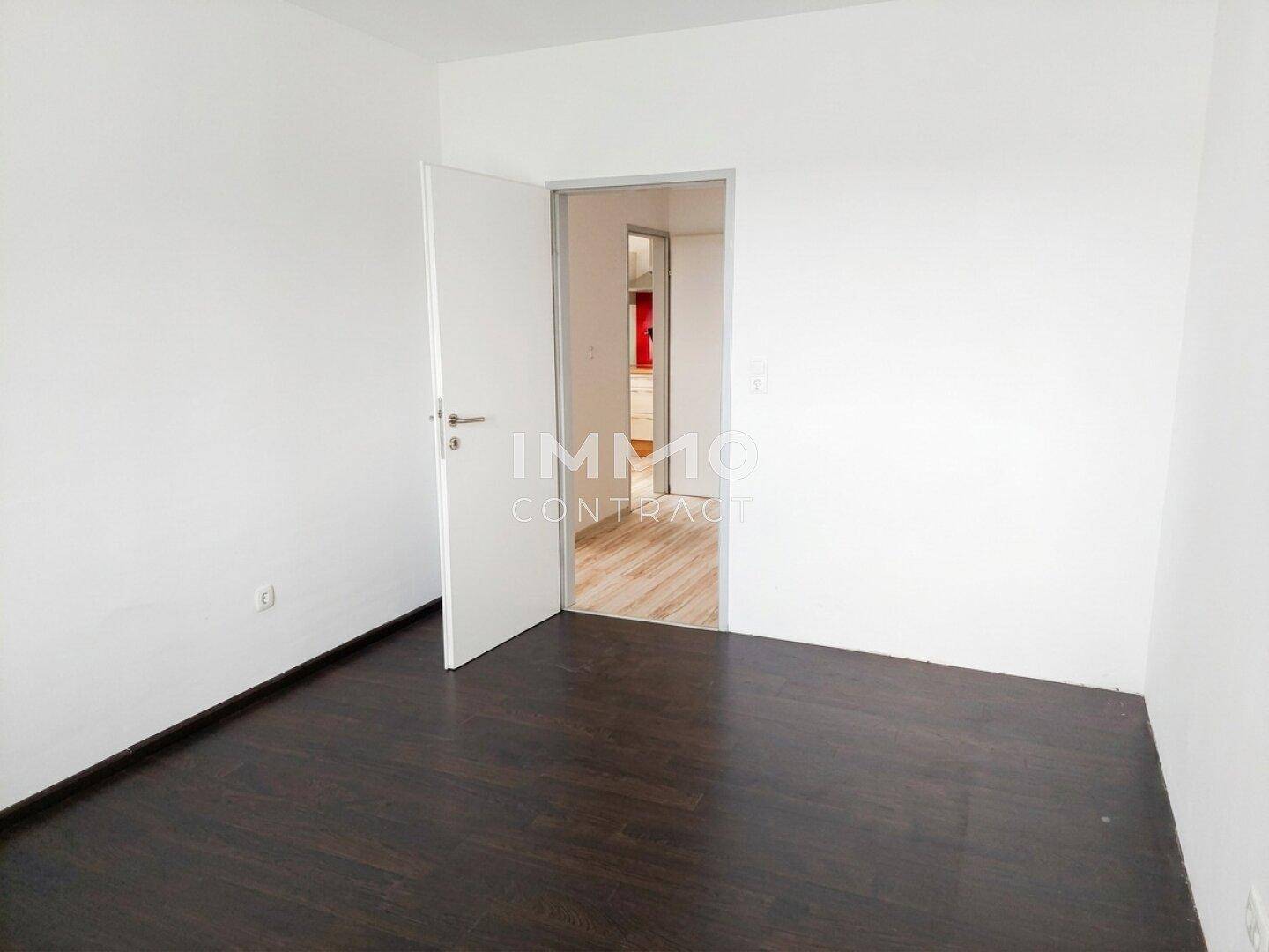 Helles Schlafzimmer mit dunklem Laminatboden und Zugang zu einem weiteren Raum.