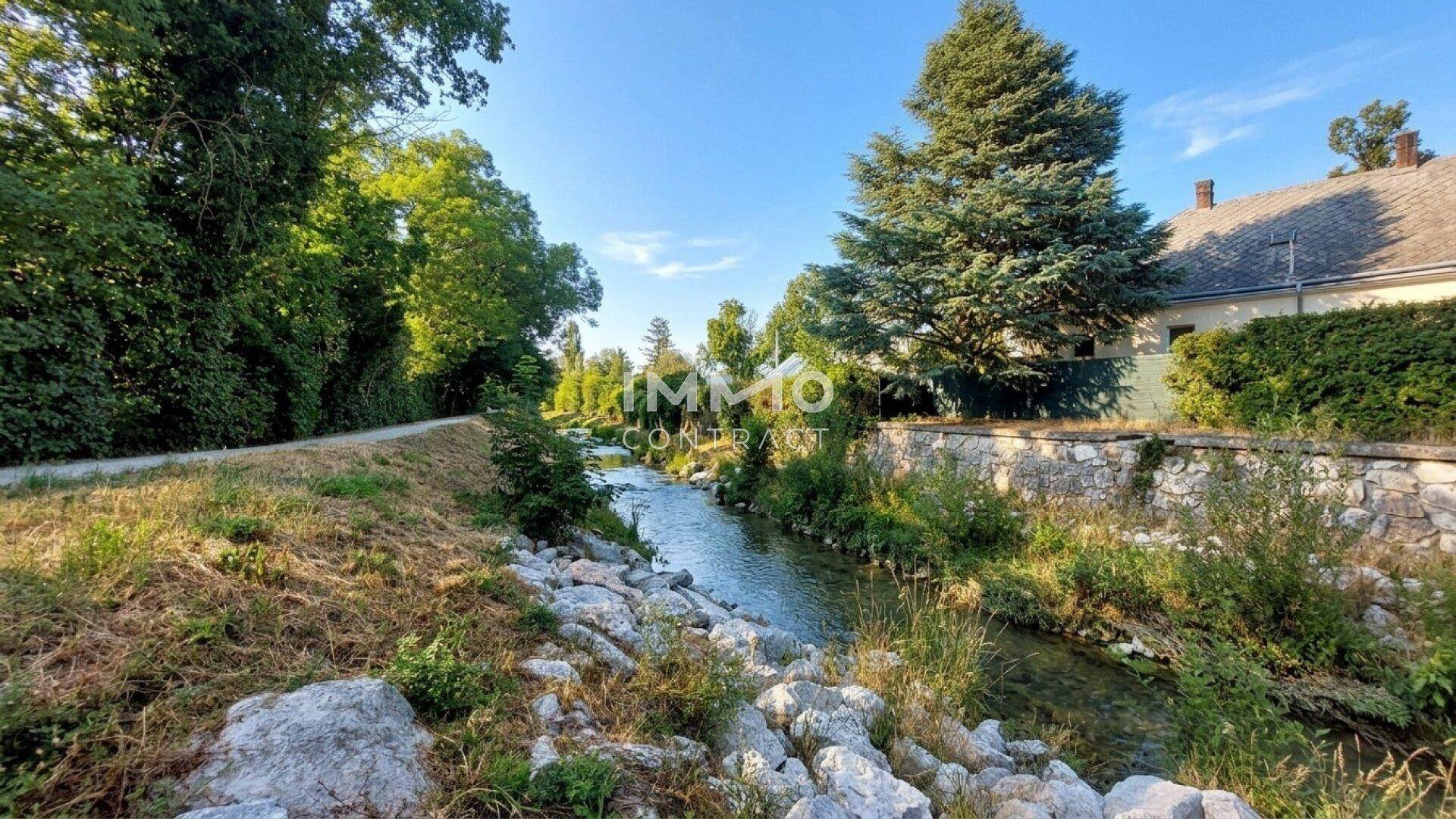 Malerischer Flusslauf mit üppiger Vegetation und einem Haus im Hintergrund an einem sonnigen Tag.