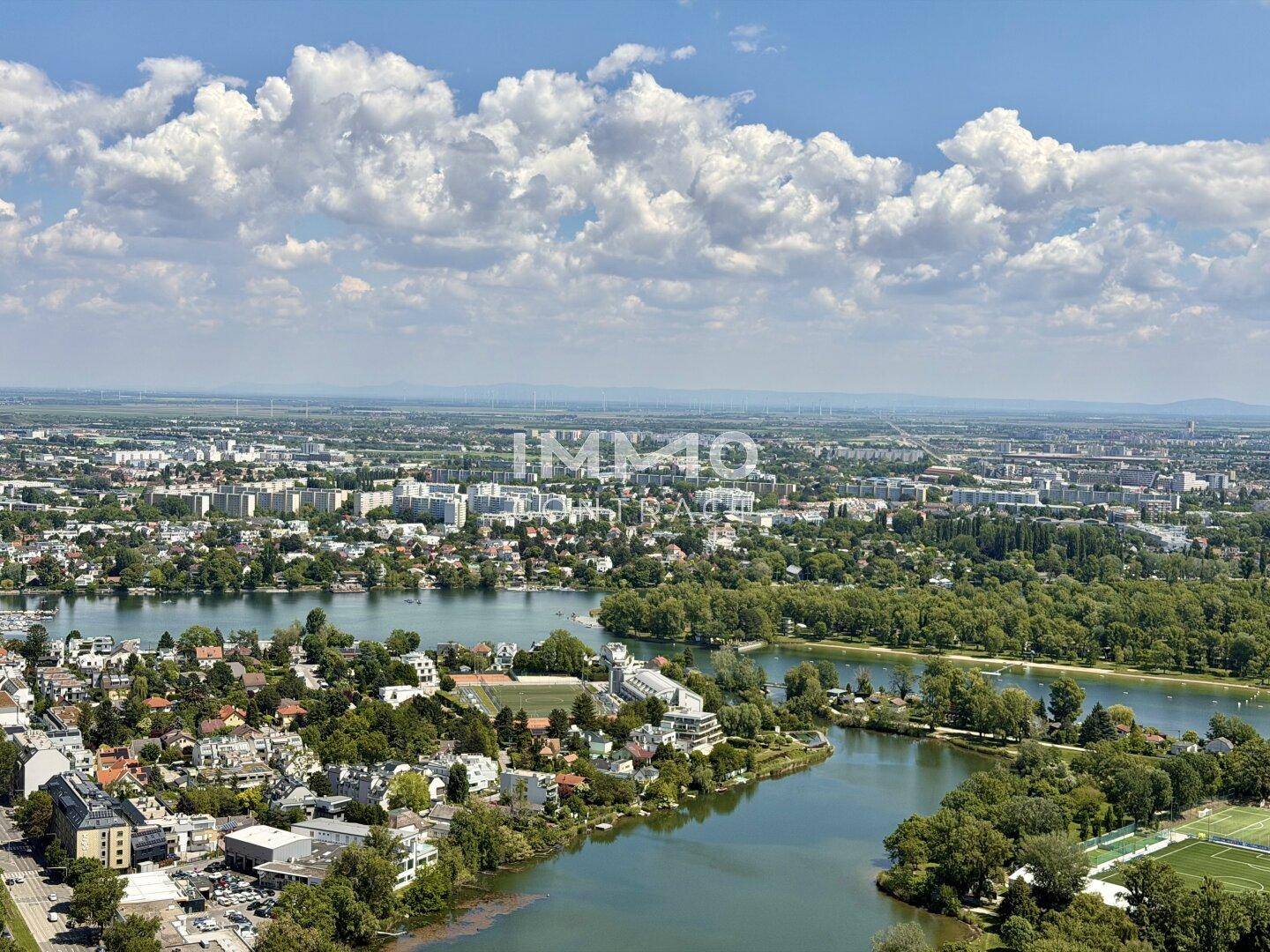 Weitläufiger Blick über die Stadtlandschaft mit Seen, Grünflächen und Windrädern am Horizont.