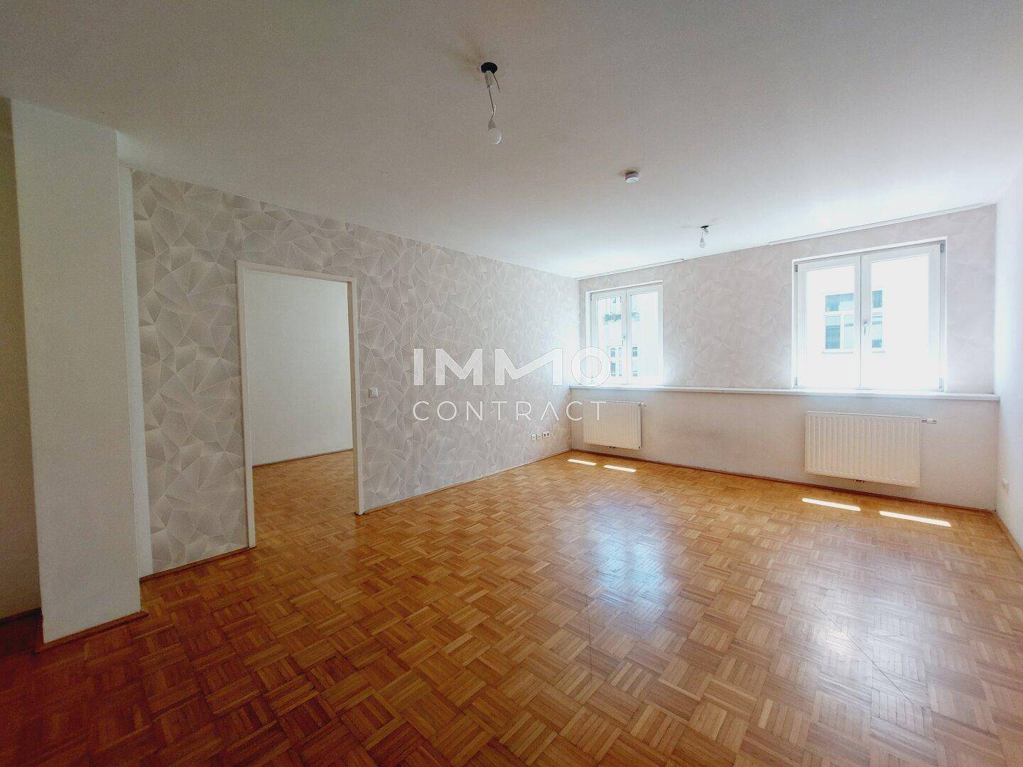 Geräumiges Wohnzimmer mit Parkettboden, gemusterter Tapete und zwei großen Fenstern.