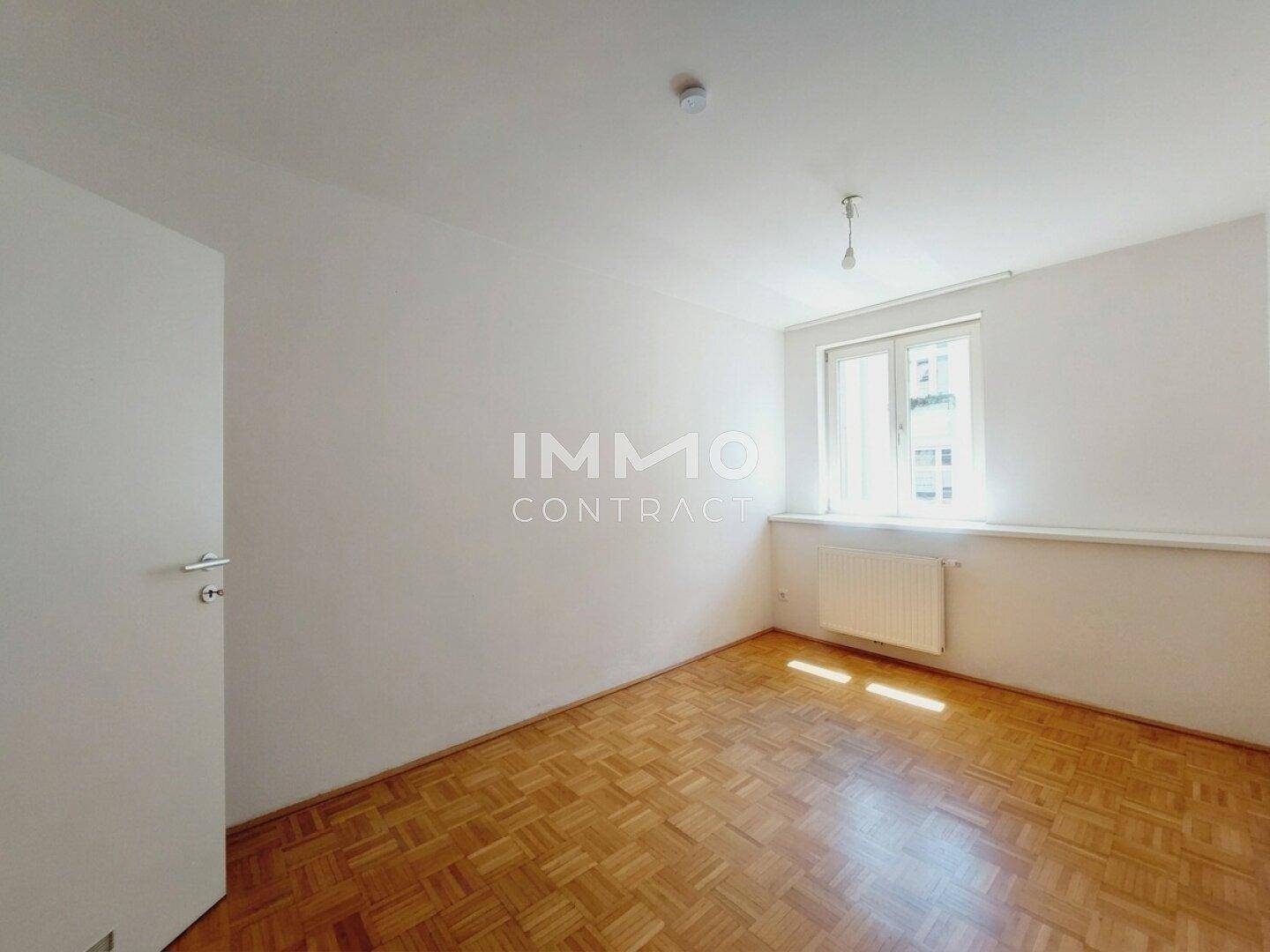 Helles Schlafzimmer mit Parkettboden, großem Fenster und Heizkörper unter der Fensterbank.
