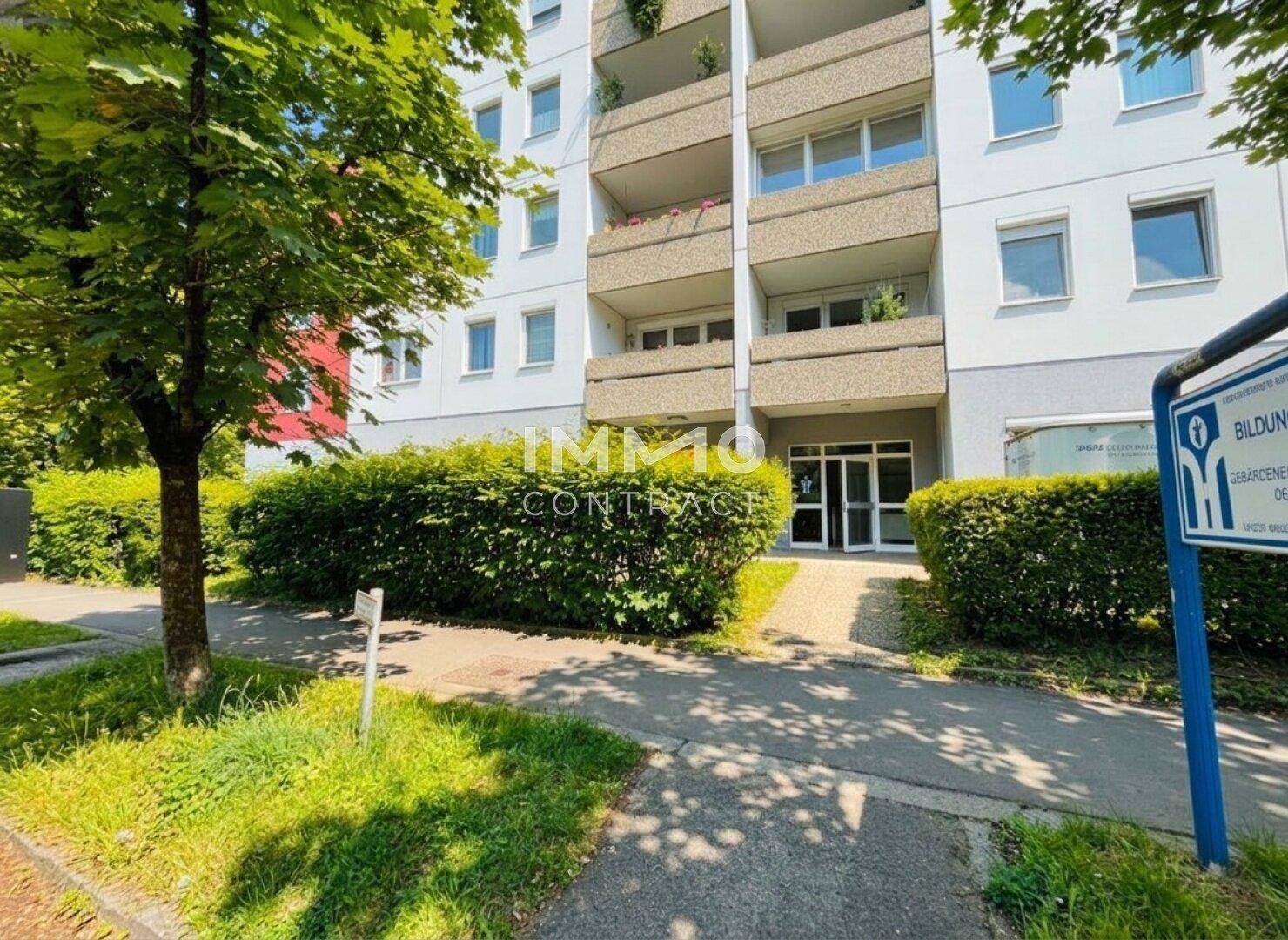 Ansicht eines Mehrfamilienhauses mit mehreren Balkonen und grünen Hecken entlang des Gehwegs.