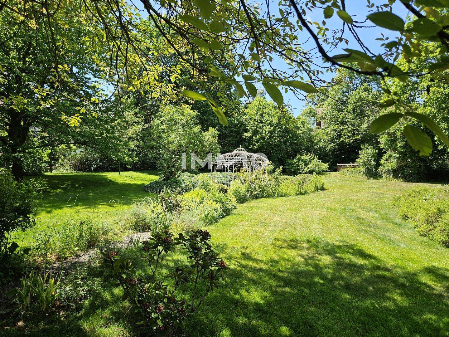 Ruhiger Gartenbereich mit üppiger Vegetation, altem Baumbestand und einem charmanten Pavillon im Hintergrund.