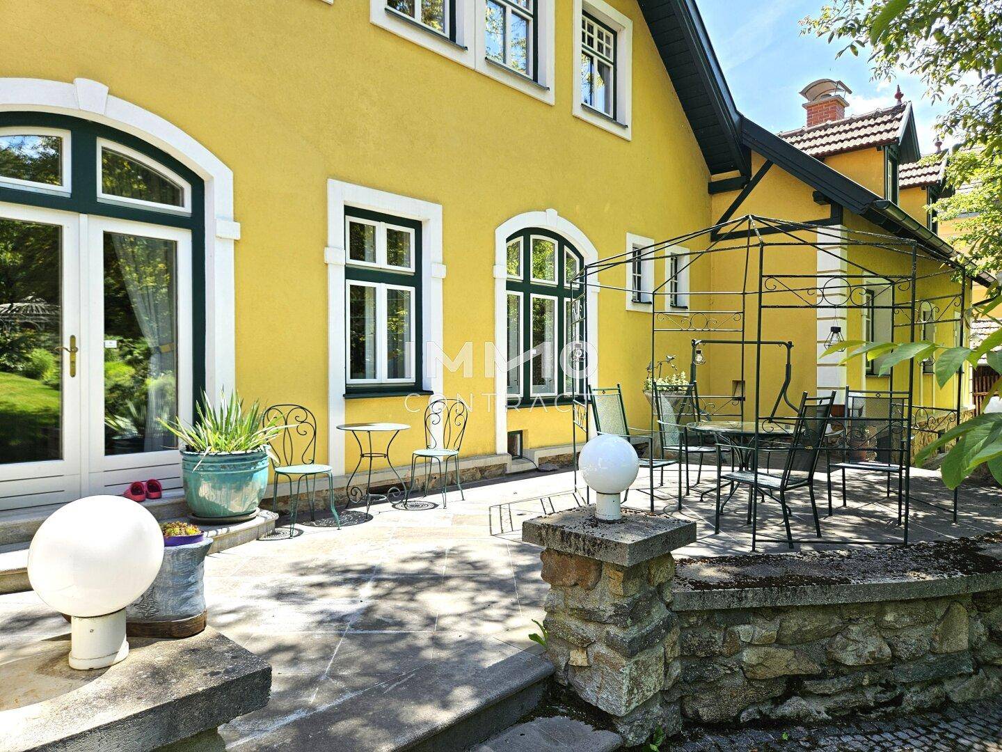 Geräumige Steinterrasse mit Gartenmöbeln und einem schmiedeeisernen Pavillon, umgeben von Grün.