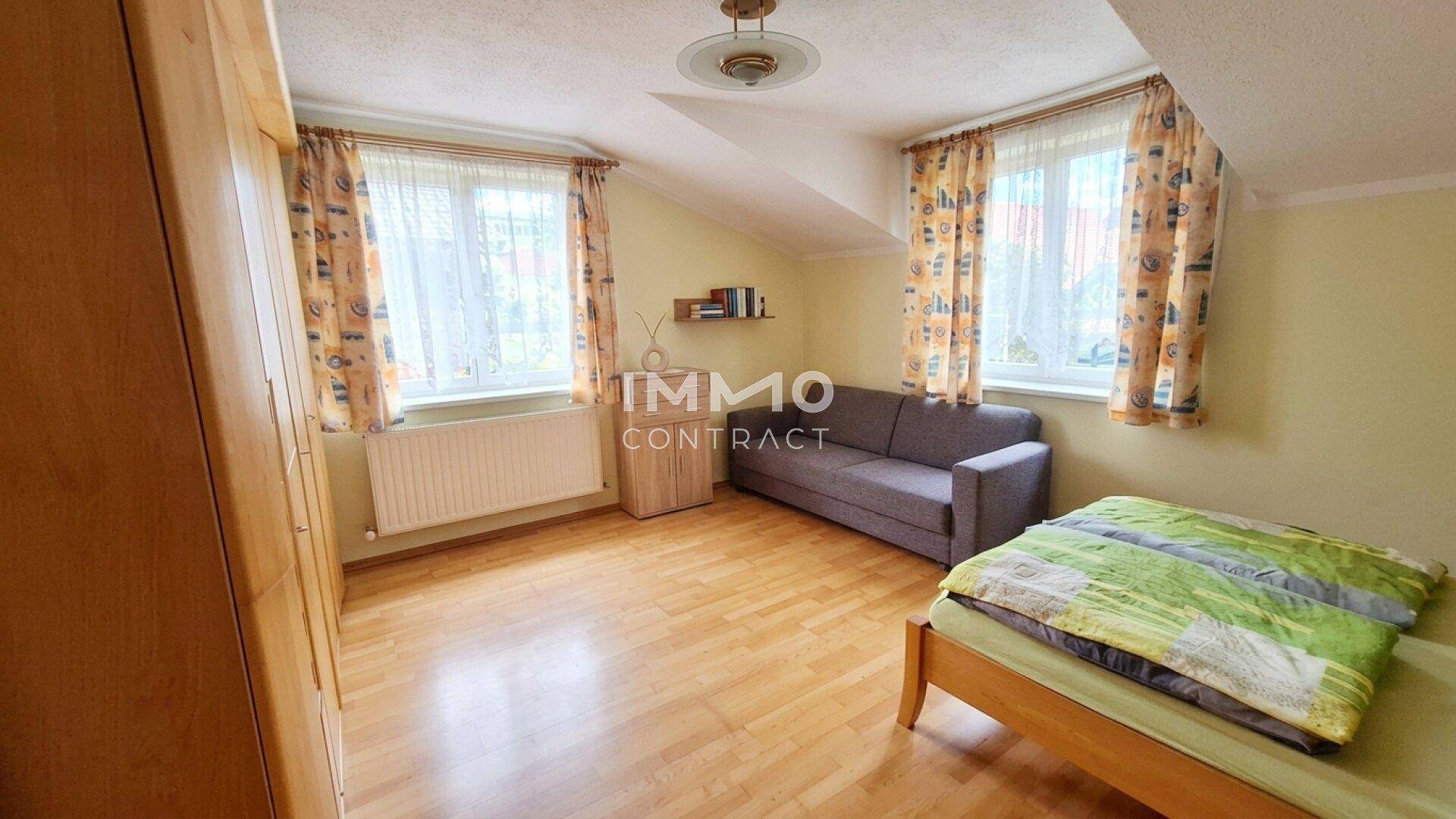Schlafzimmer mit Holzboden, zwei Fenstern und einer Dachschräge, ausgestattet mit einem Bett und Schrank.