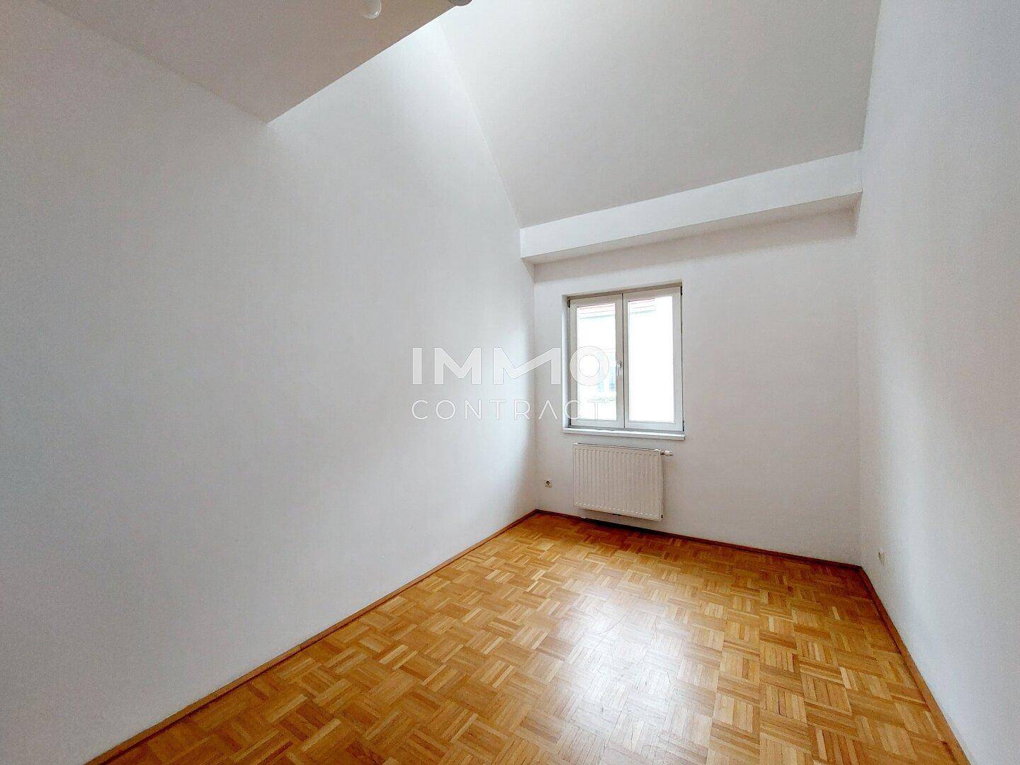 Helles Schlafzimmer mit Parkettboden, einem Fenster und schräger Decke.