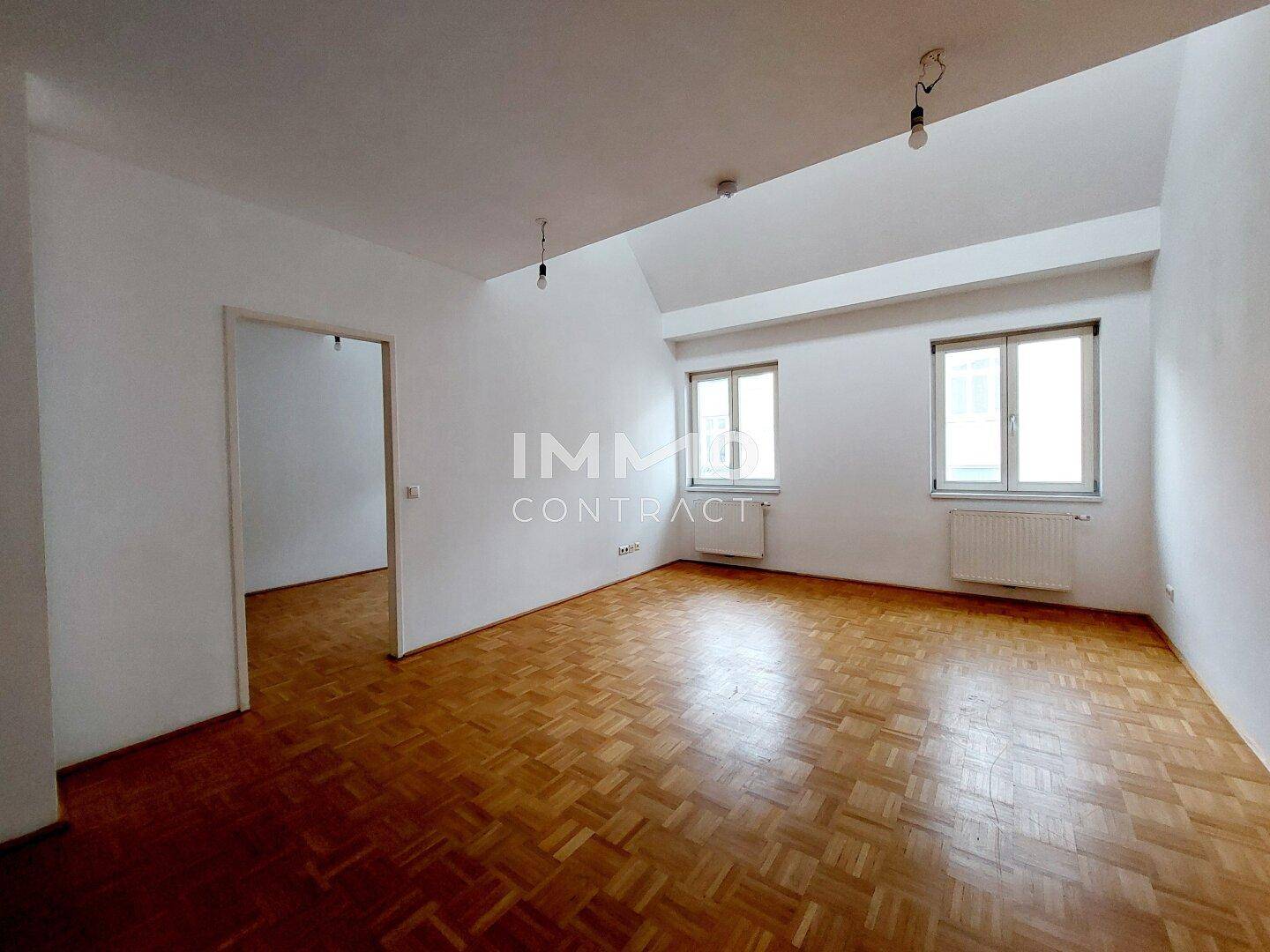 Geräumiges Wohnzimmer mit Parkettboden, zwei Fenstern und schrägen Decken.
