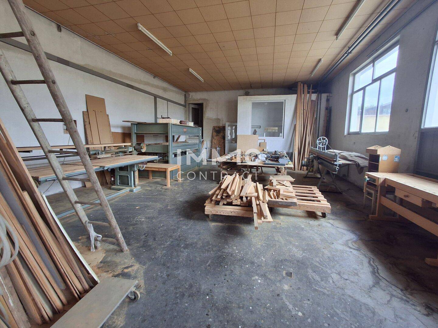 Großer Werkstattraum mit Holzmaterialien, Werkbänken und Tageslicht durch Fenster.
