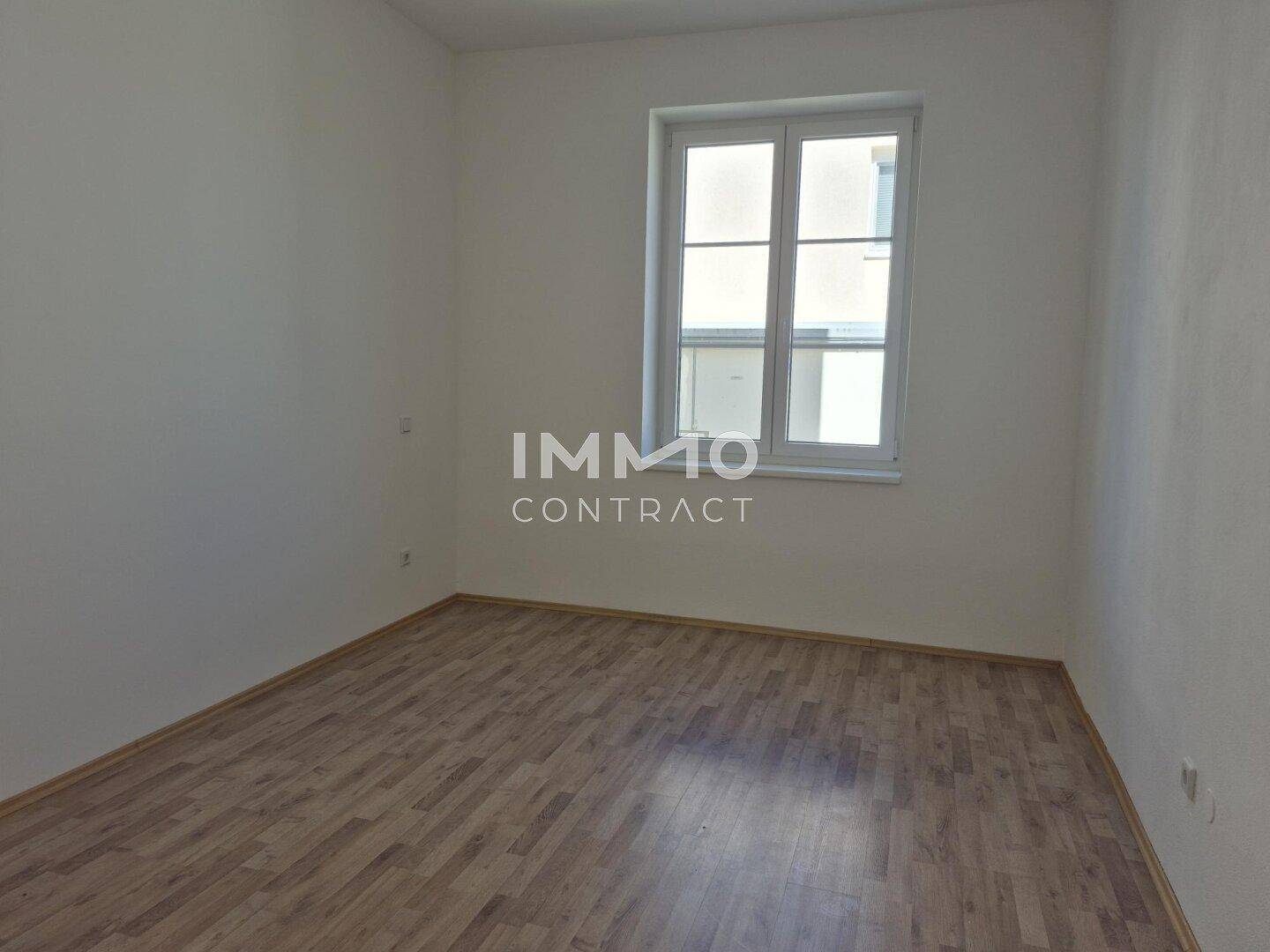 Leeres Zimmer mit Laminatboden und einem Fenster, das viel Tageslicht hereinlässt.