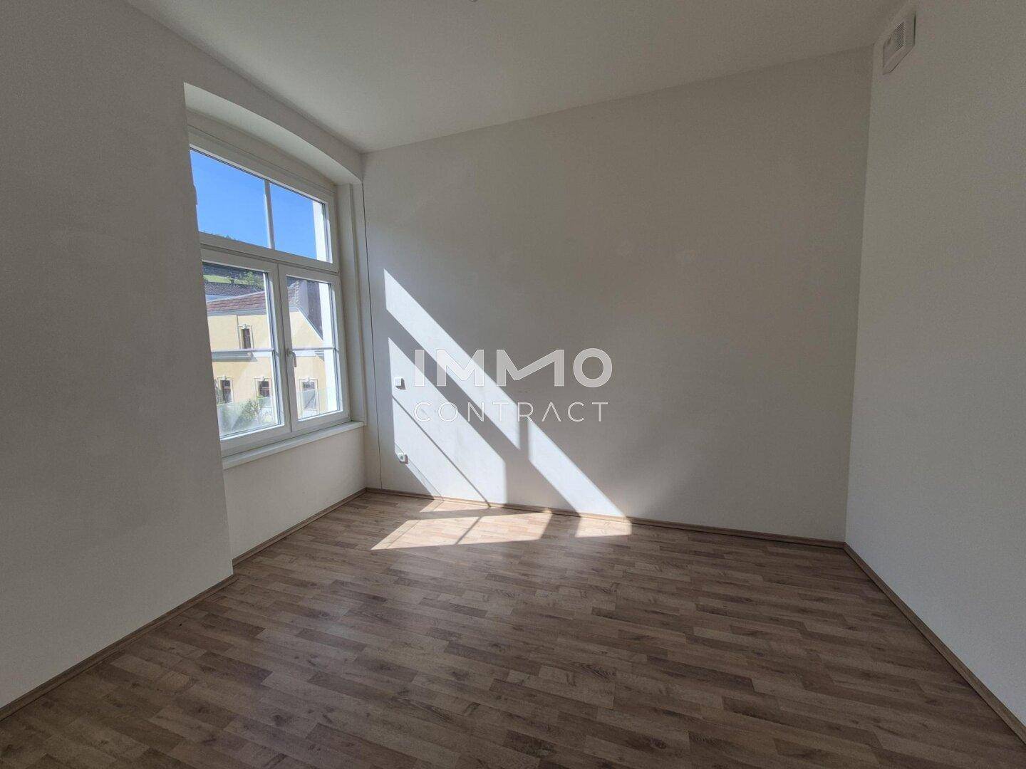 Heller Raum mit Holzboden und einem großen Fenster, das viel Sonnenlicht hereinlässt.