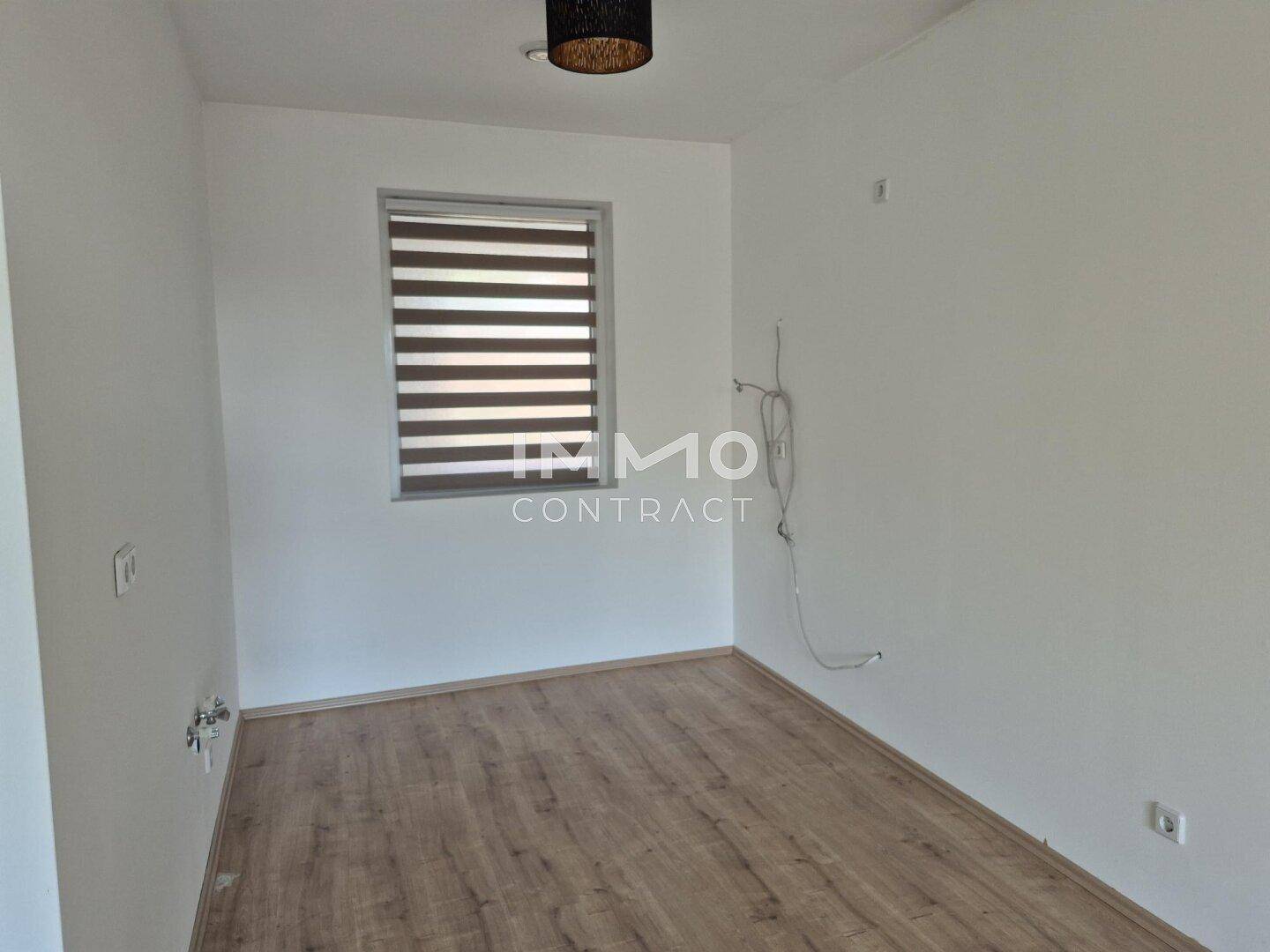 Leeres Zimmer mit Laminatboden, weißen Wänden und einem Fenster mit Jalousien.