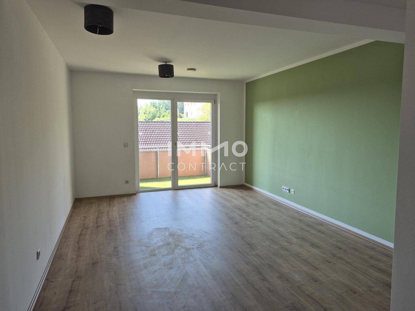 Geräumiges Wohnzimmer mit Laminatboden, grüner Akzentwand und Zugang zu einem Außenbereich.