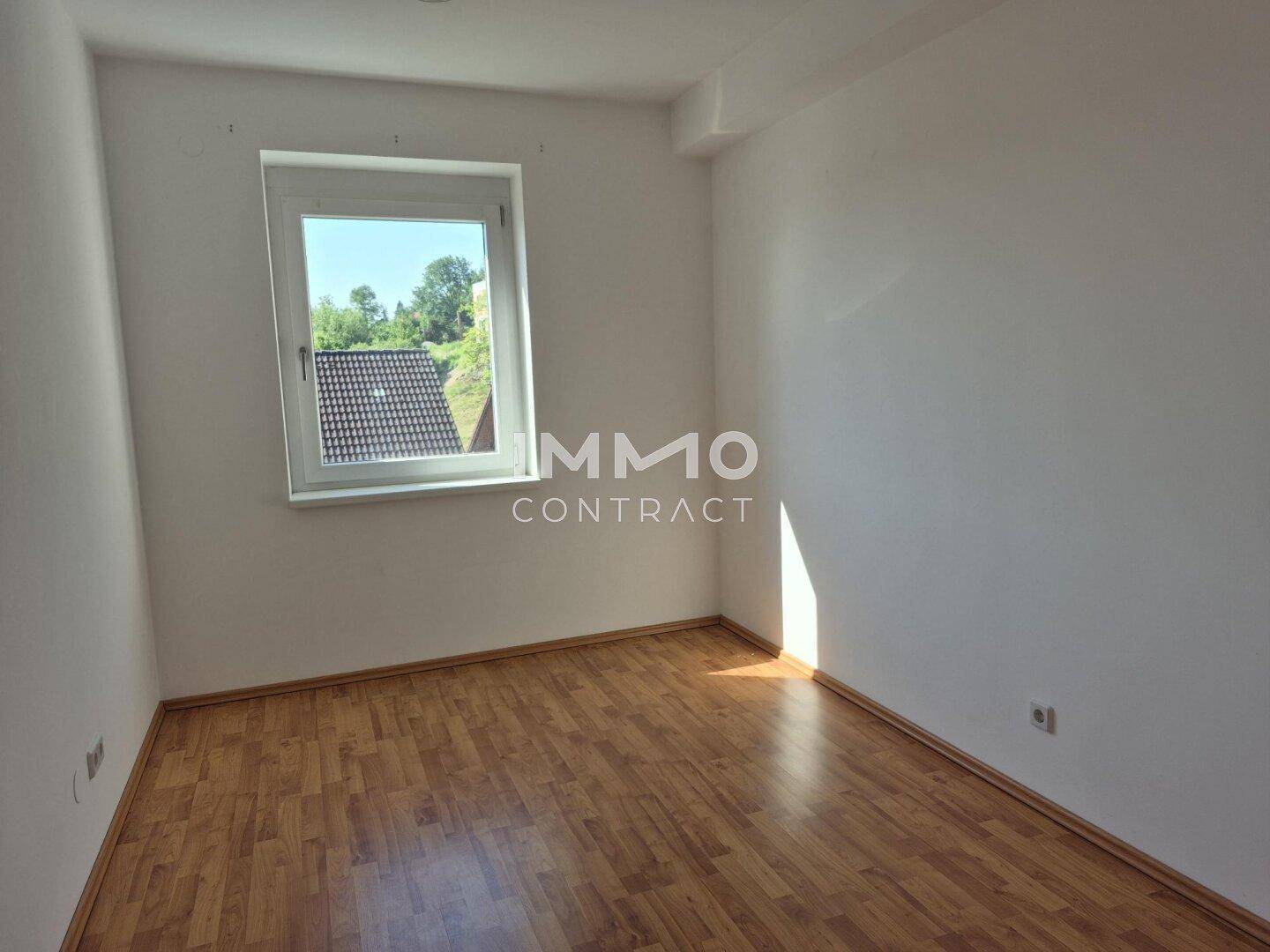 Helles Zimmer mit Laminatboden, weißen Wänden und Blick auf Dächer und Grünflächen.