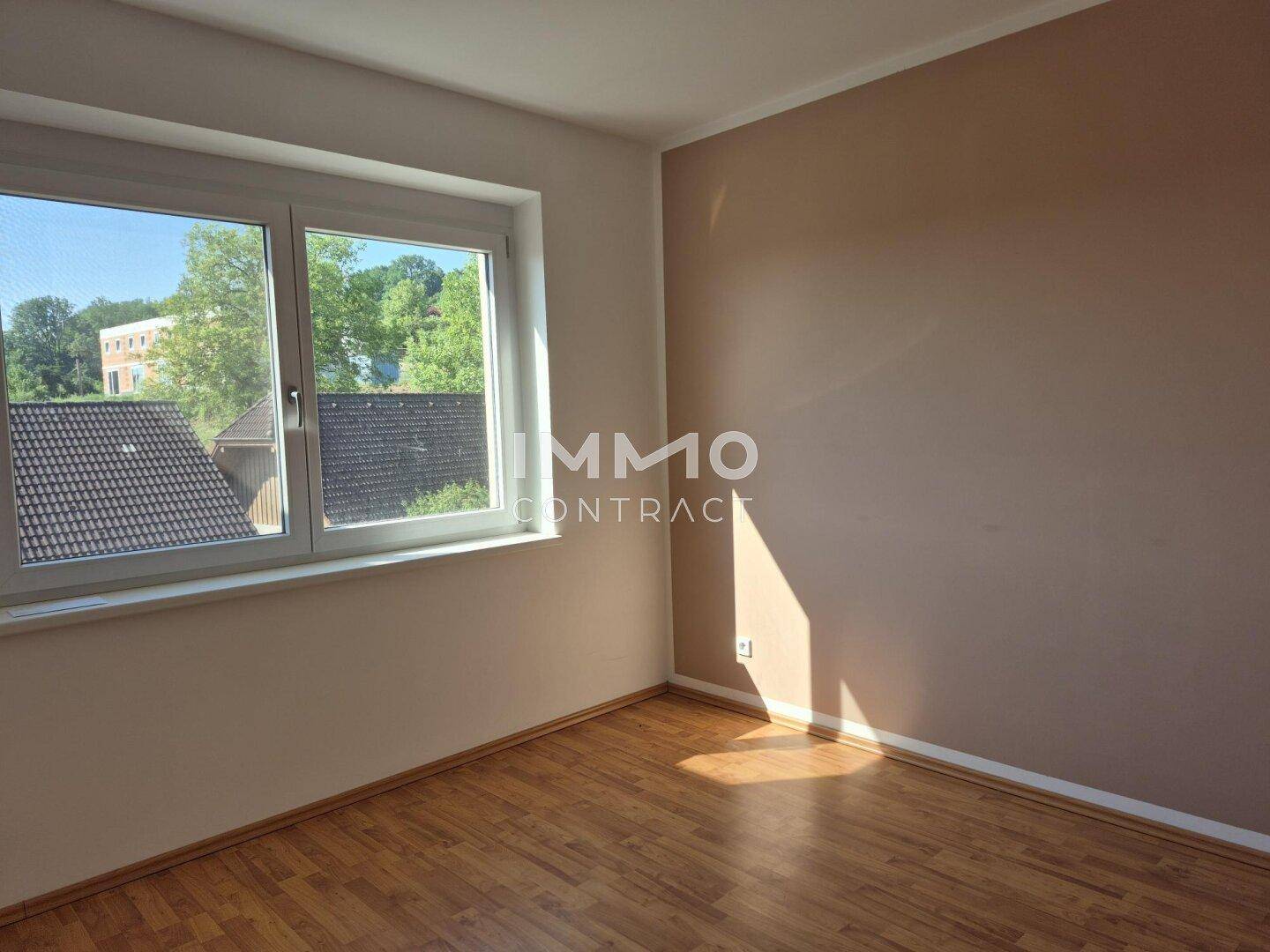 Geräumiges Zimmer mit Holzboden, hellbraunen Wänden und weitem Blick ins Grüne.