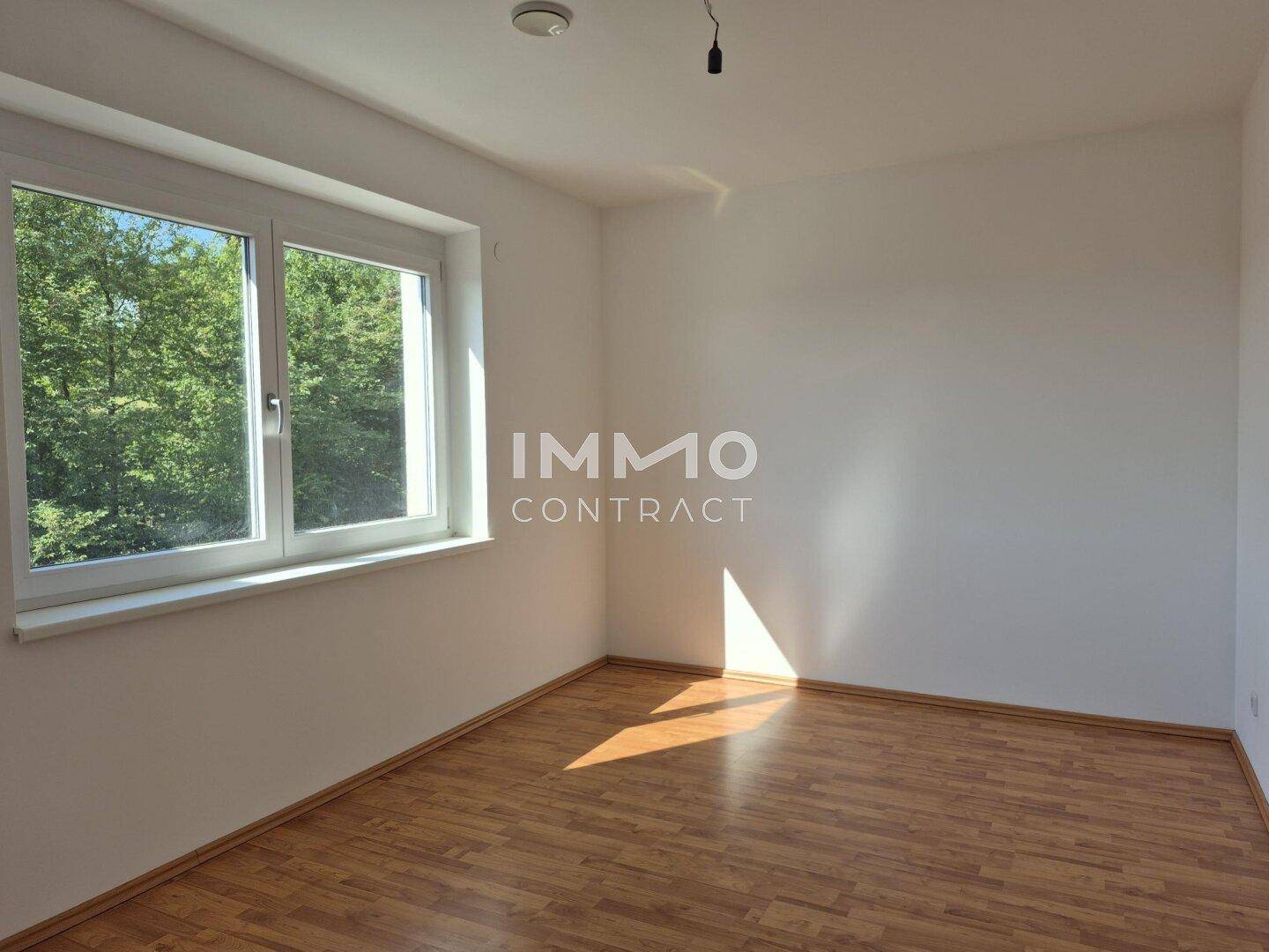 Helles Zimmer mit großem Fenster, Blick ins Grüne und Sonnenlicht auf dem Boden.