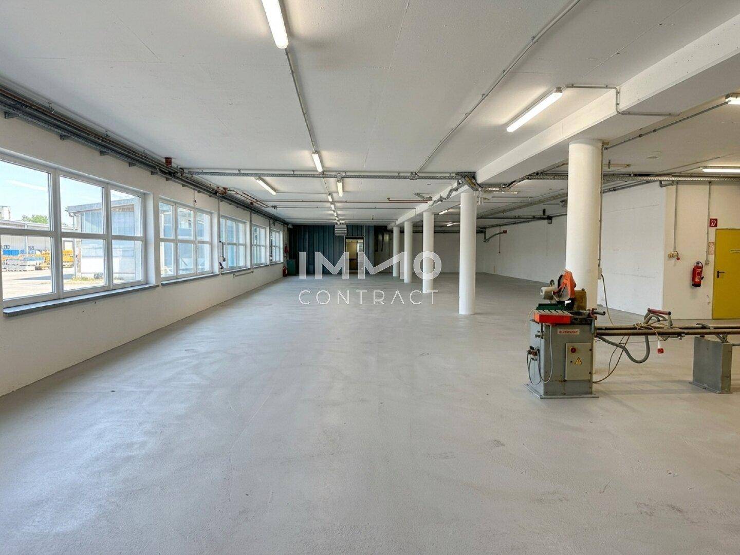 Weitläufige Lagerhalle mit Betonboden, Säulen und großen Fenstern, teilweise mit Maschinen.