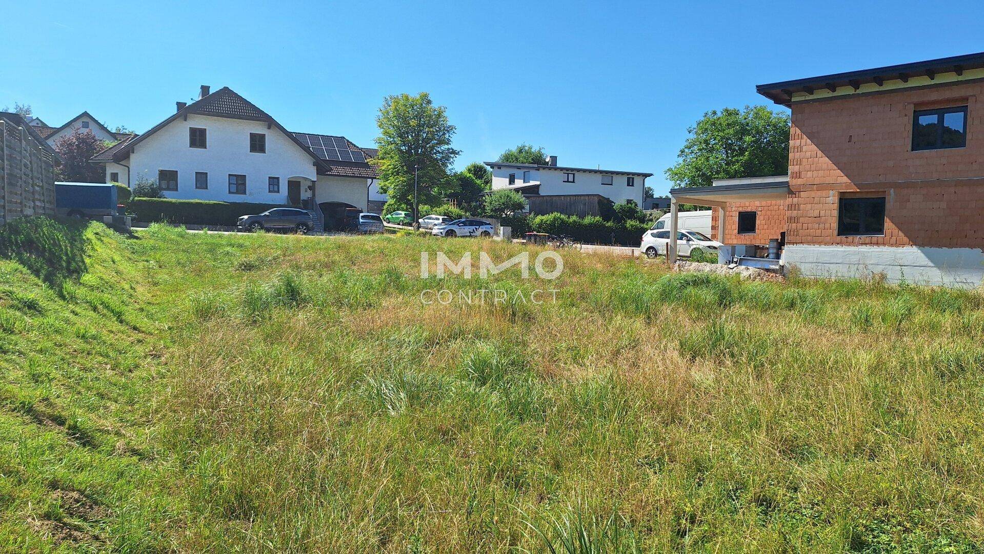 Großes Grundstück mit Wiese und einem im Bau befindlichen Haus, umgeben von weiteren Wohnhäusern an einem sonnigen Tag.