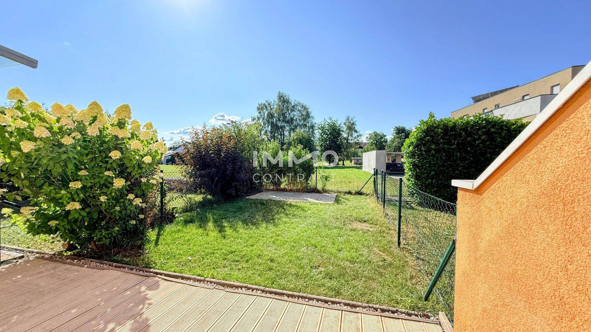 Gepflegter Gartenbereich mit Rasenfläche, Sträuchern und einer Holzterrasse im Vordergrund.