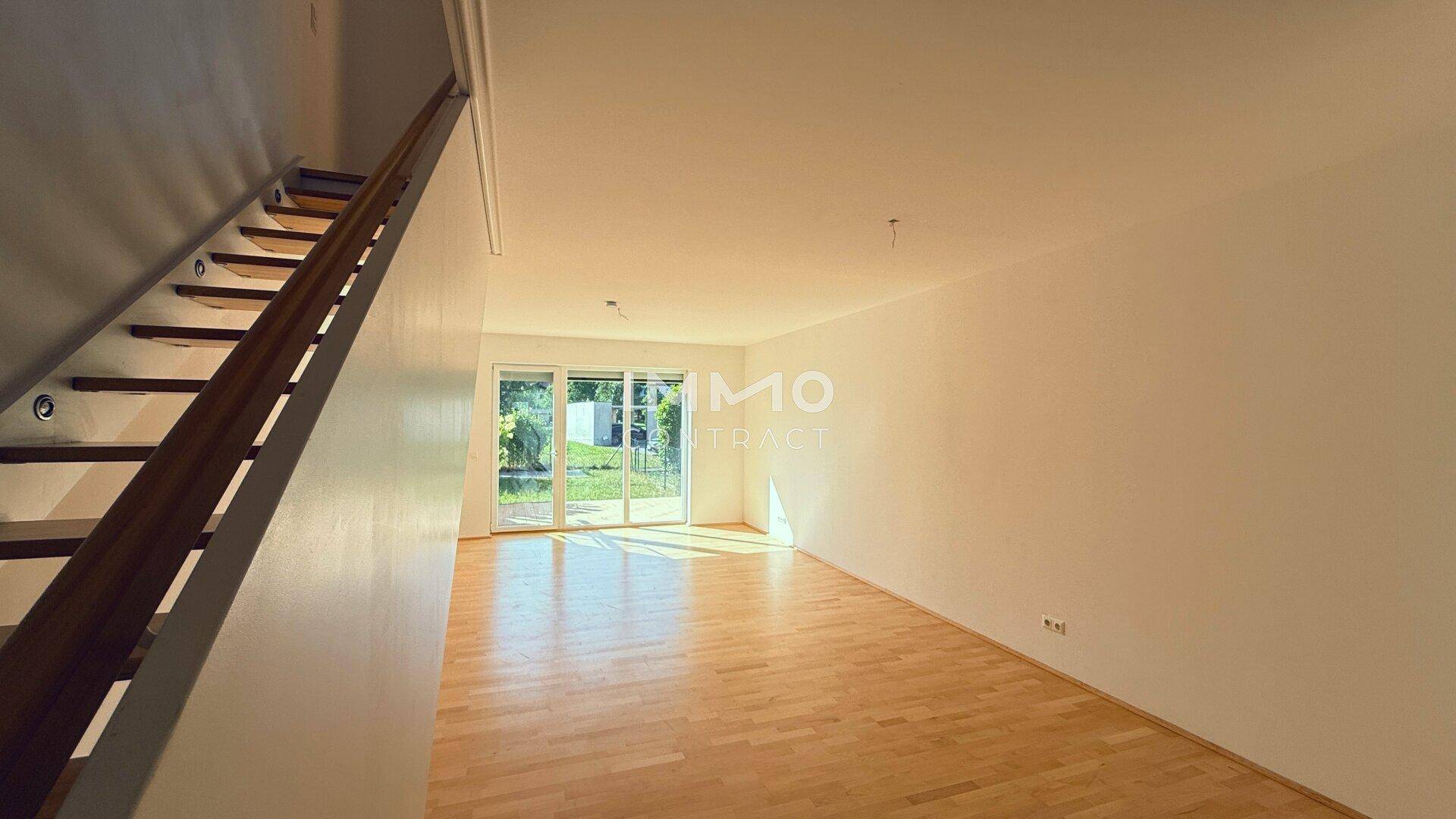 Helles, leeres Wohnzimmer mit Holzfußboden und großem Zugang zum Gartenbereich.