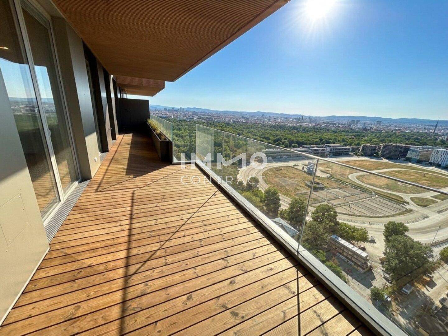 Großer Holzbalkon mit Glasgeländer und weitem Blick über die Stadt und grüne Flächen.