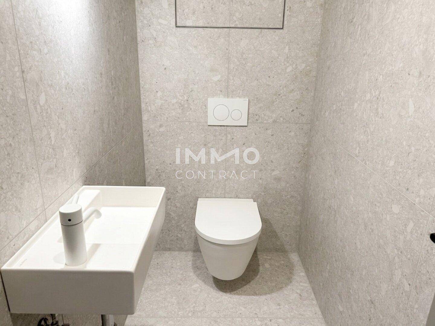Modernes Gäste-WC mit grauen Fliesen, wandhängender Toilette und minimalistischem Waschbecken.