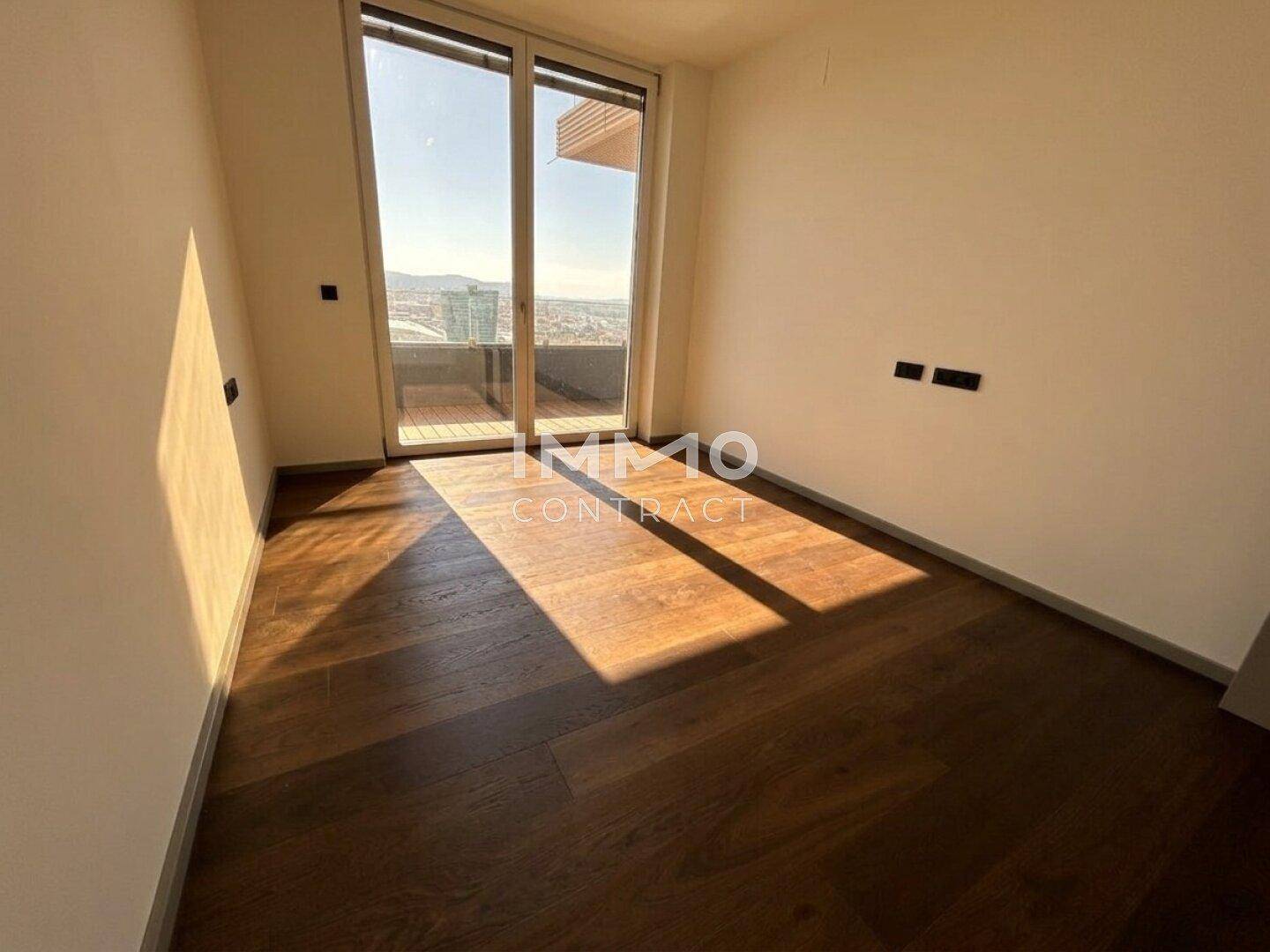 Sonniger Raum mit Holzboden und großen Fenstern, die Zugang zu einem Außenbereich bieten.