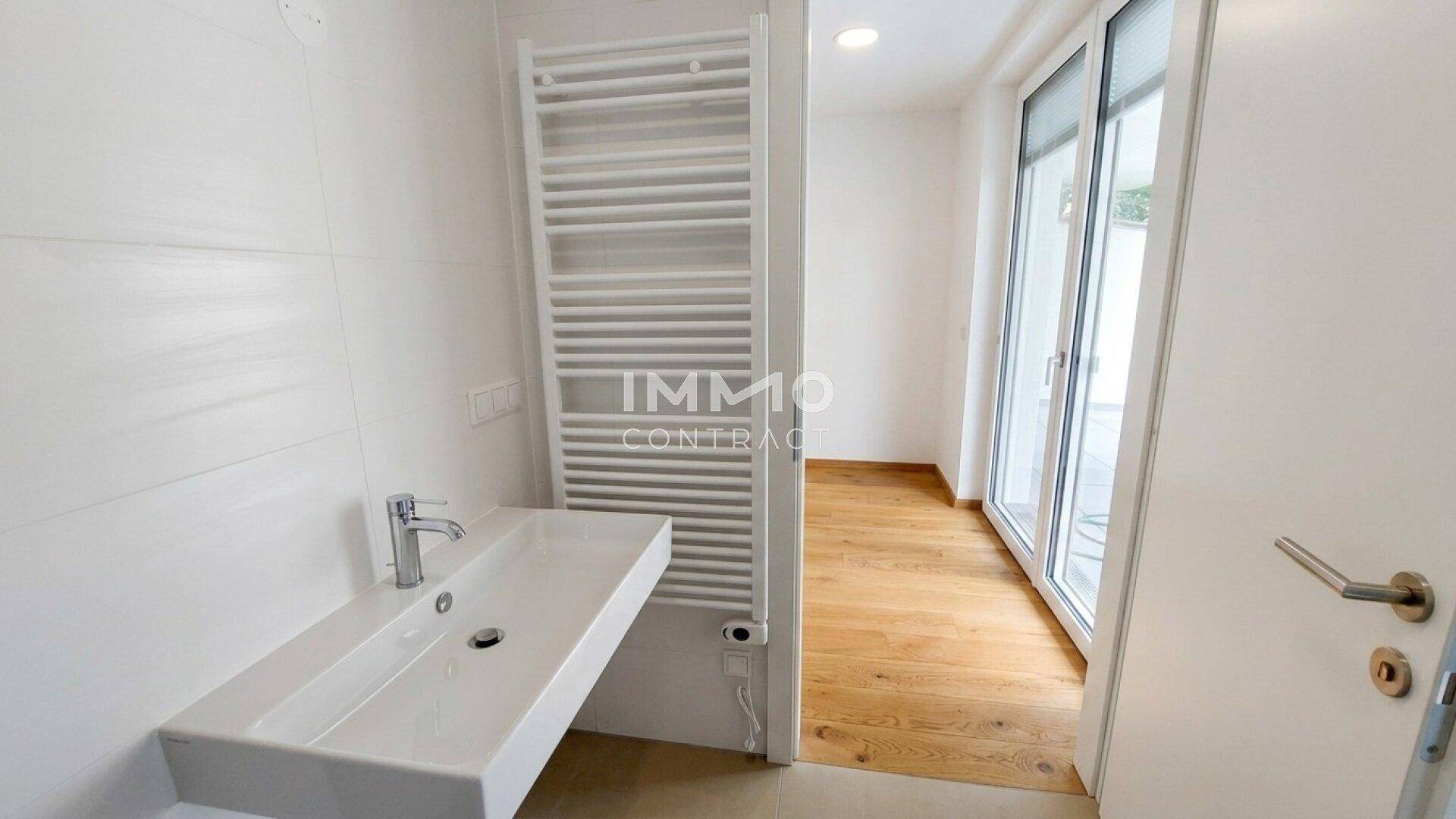 Helles Badezimmer mit Waschbecken, beheiztem Handtuchhalter und Zugang zu einem weiteren Raum.