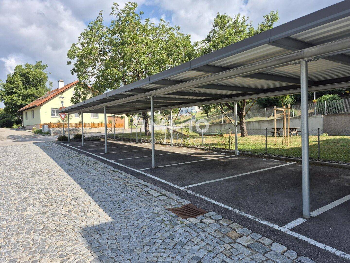 Überdachte Parkplätze (Carport) auf einem gepflasterten Bereich neben einem Wohngebäude.