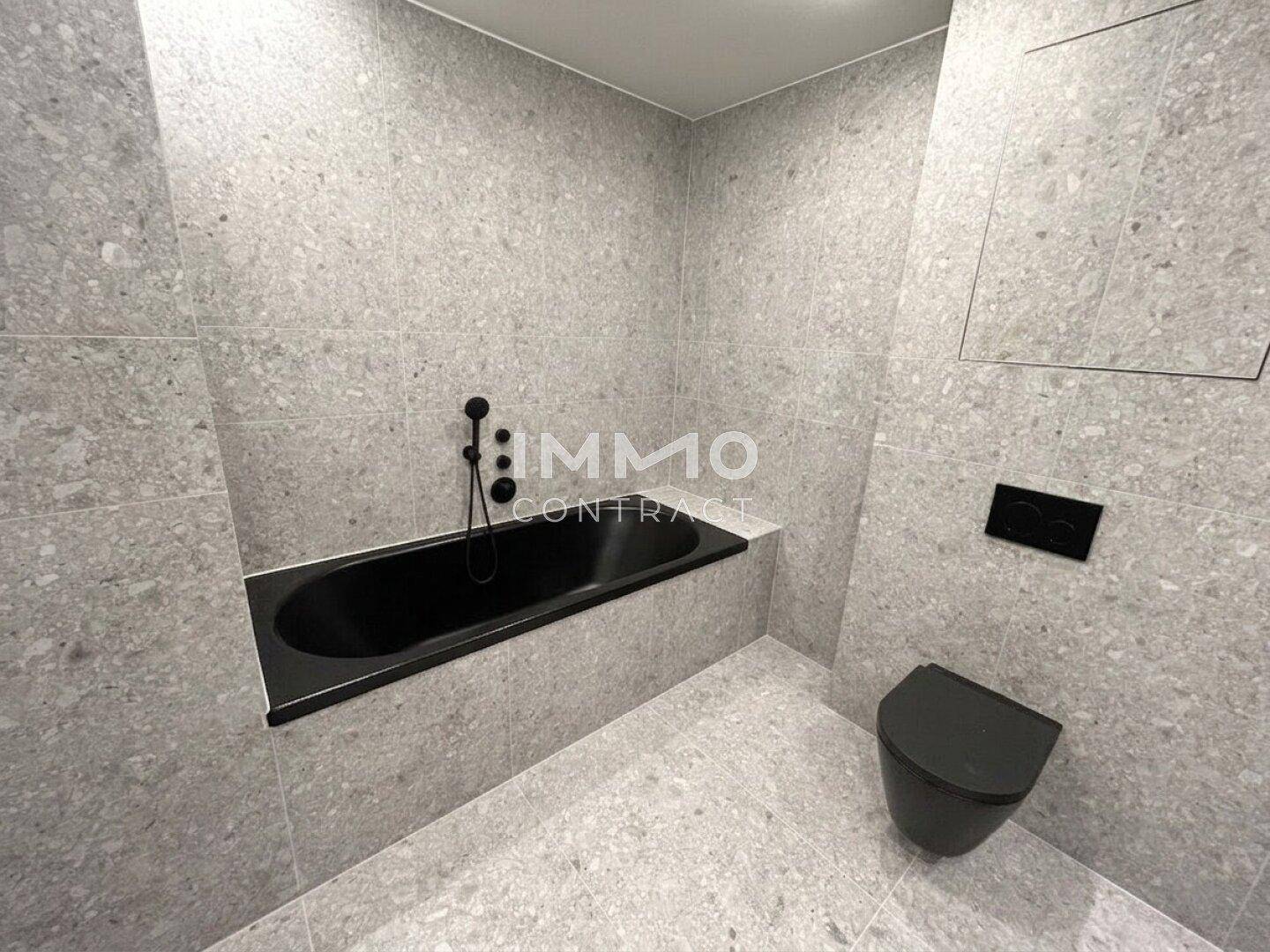 Modernes Badezimmer mit integrierter Badewanne, grauen Fliesen und schwarzer Toilette.