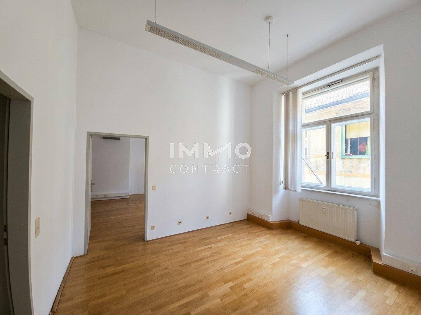 Heller Raum mit Holzboden, großem Fenster und Zugang zu einem weiteren Zimmer.