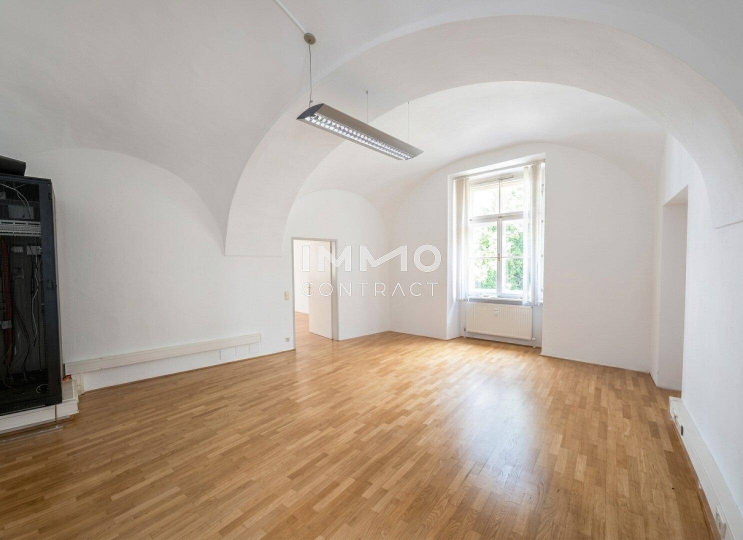 Großer Raum mit eleganter Gewölbedecke, Holzboden und viel Tageslicht durch das Fenster.