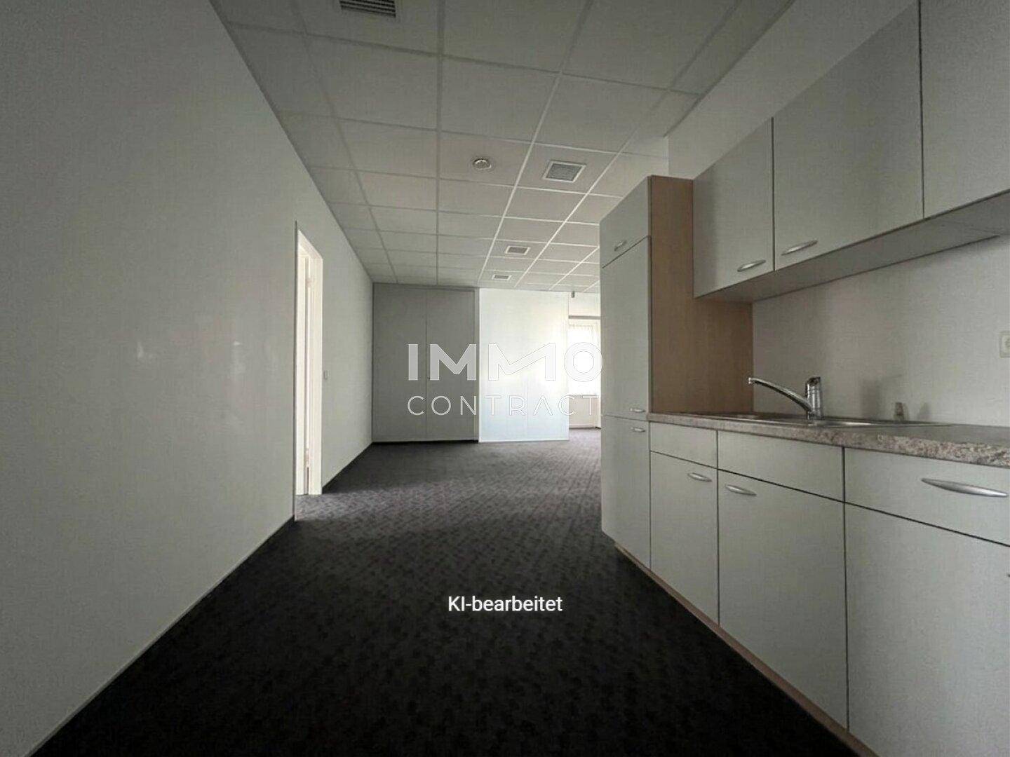 Leeres Büro mit dunklem Teppichboden, abgehängter Decke und einer funktionalen Küchenzeile.