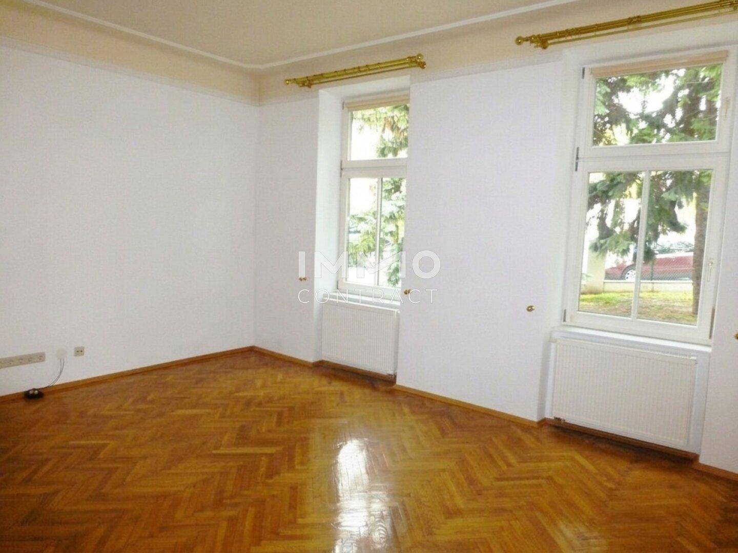Leeres Zimmer mit Fischgrätparkett, weißen Wänden und zwei großen Fenstern.