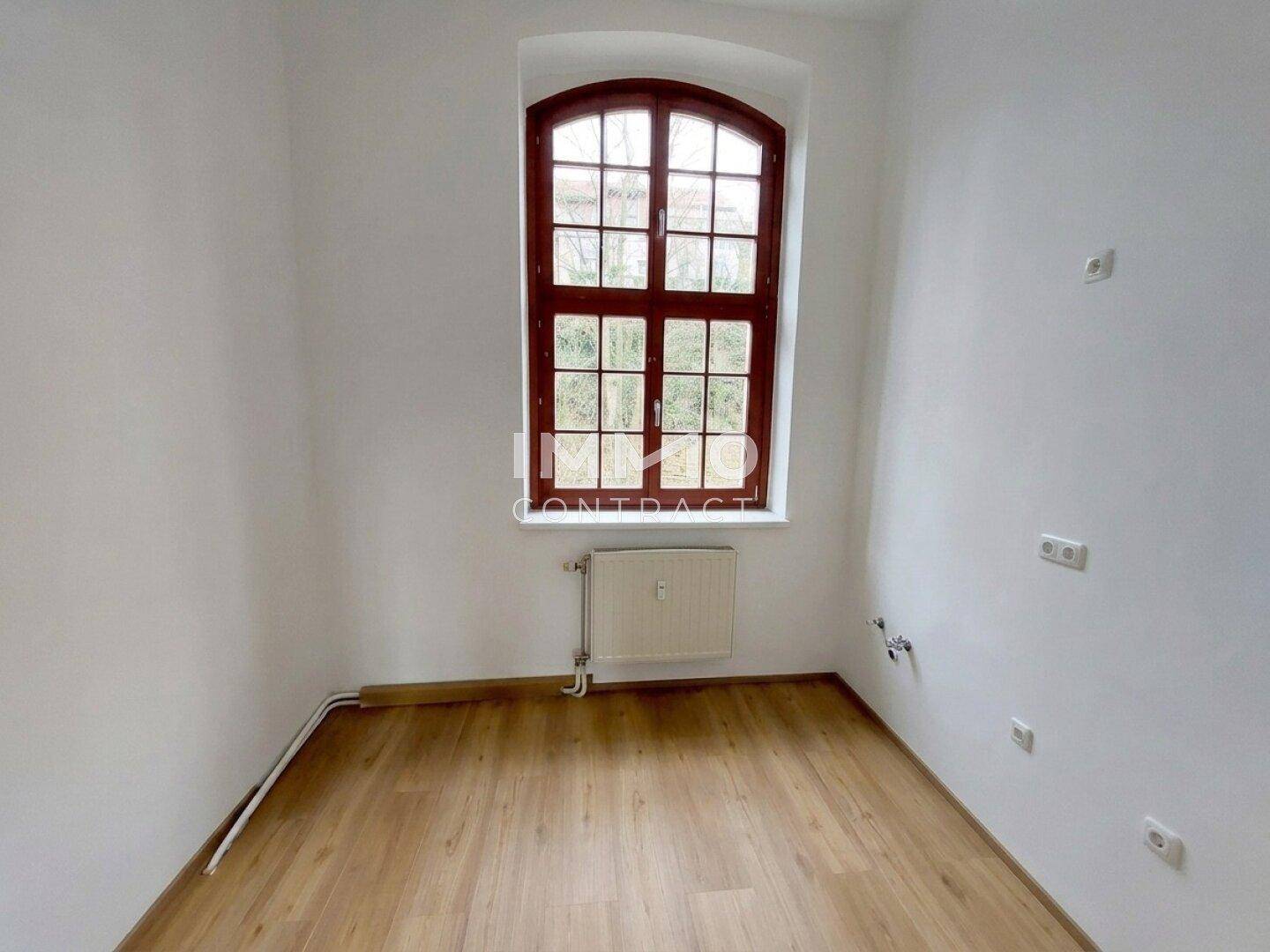 Leeres Zimmer mit Laminatboden und einem hohen Bogenfenster, das viel Licht spendet.