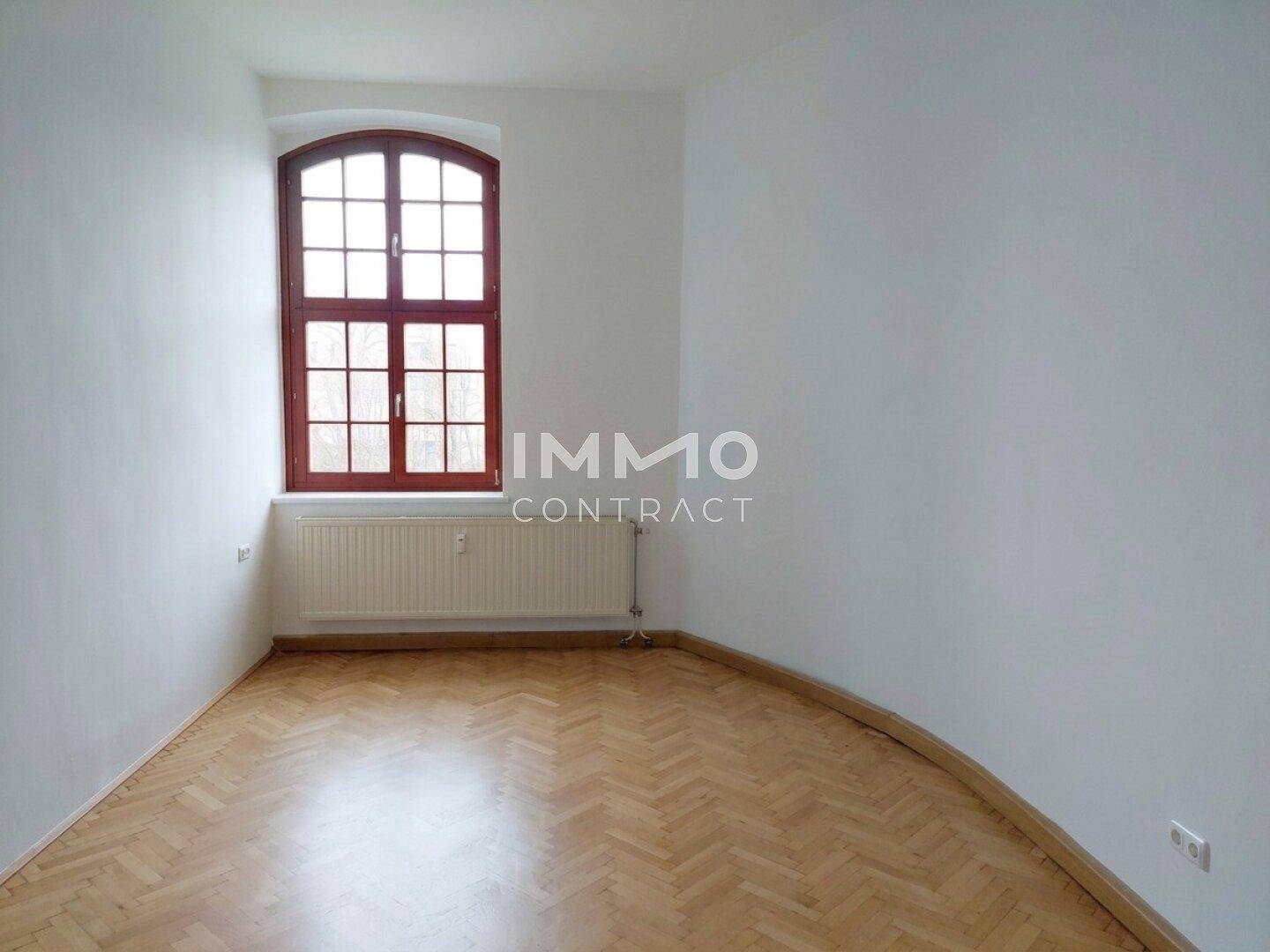 Helles Zimmer mit Parkettboden und einem hohen Bogenfenster, das eine grüne Aussicht bietet.