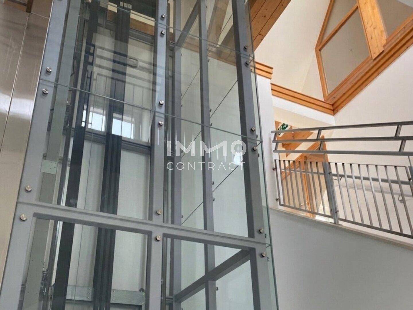 Moderner Glasaufzug in einem hellen Treppenhaus mit sichtbaren Holzbalken und Metallgeländer.