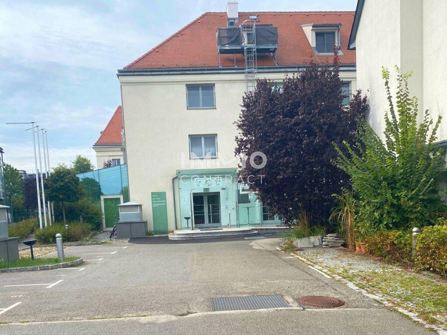 Außenansicht des Gebäudes mit Parkplatz und modernem Eingangsbereich unter bewölktem Himmel.