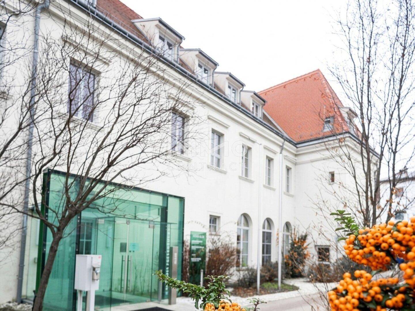 Weißes Gebäude mit rotem Ziegeldach und modernem Glaseingang, umgeben von kahlen Bäumen.