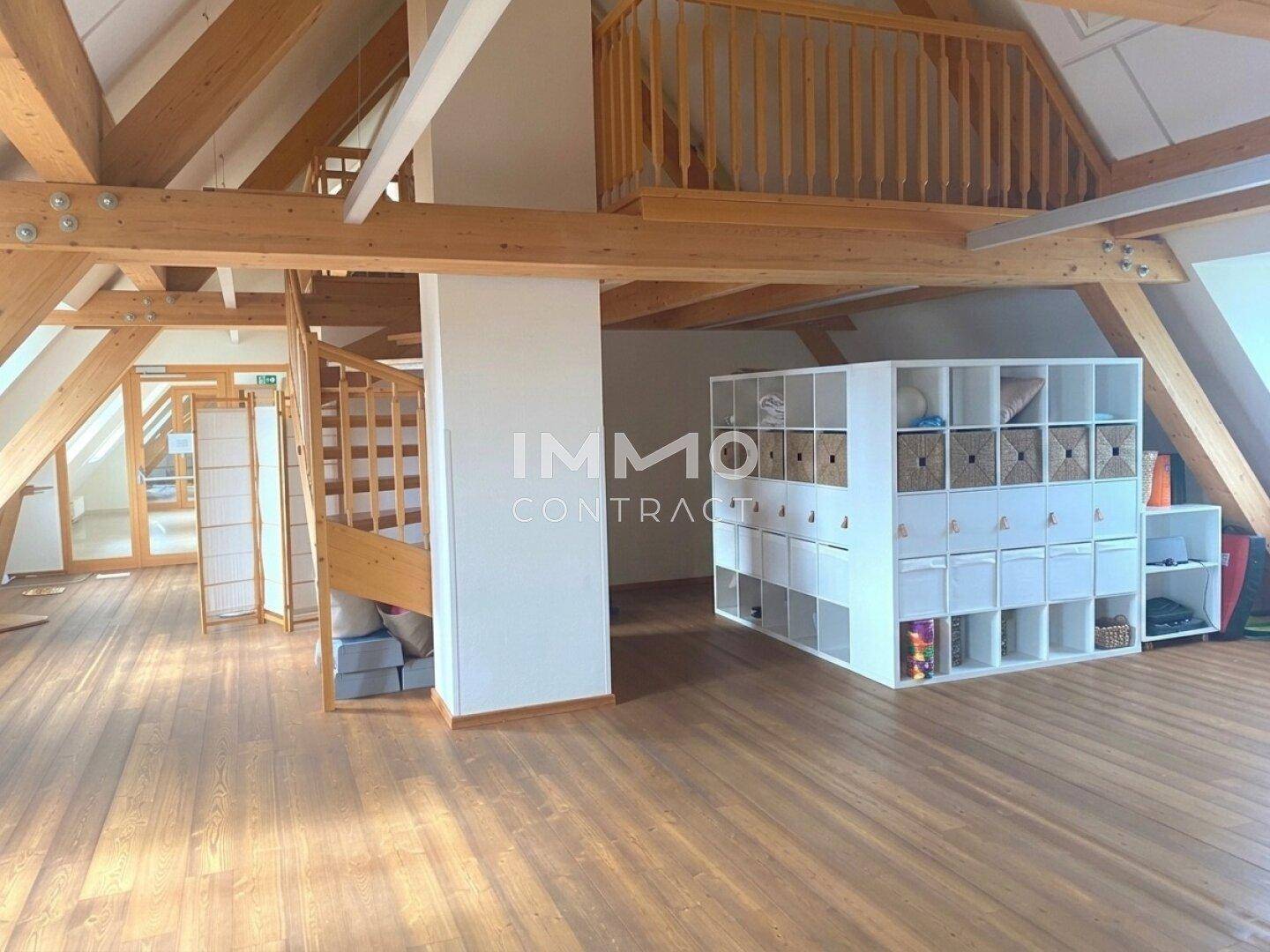 Großer offener Raum mit Holzboden, sichtbaren Dachbalken und einer Holztreppe zur Empore.