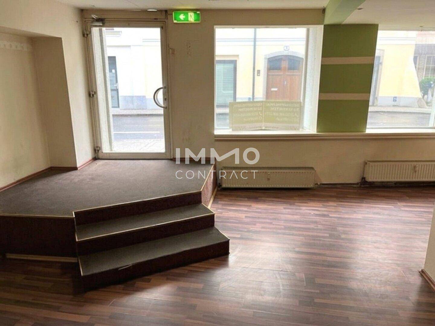 Innenansicht eines Ladenlokals mit dunklem Laminatboden, einer erhöhten Plattform und großen Schaufenstern zur Straße.