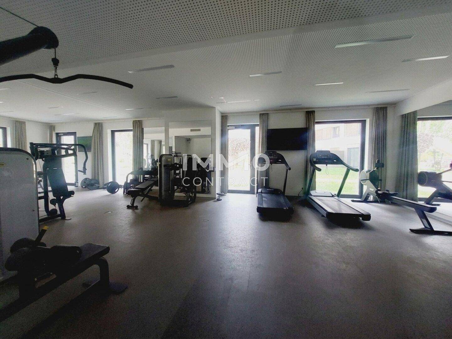 Gut ausgestatteter Fitnessraum mit verschiedenen Trainingsgeräten und großen Fenstern für Tageslicht.