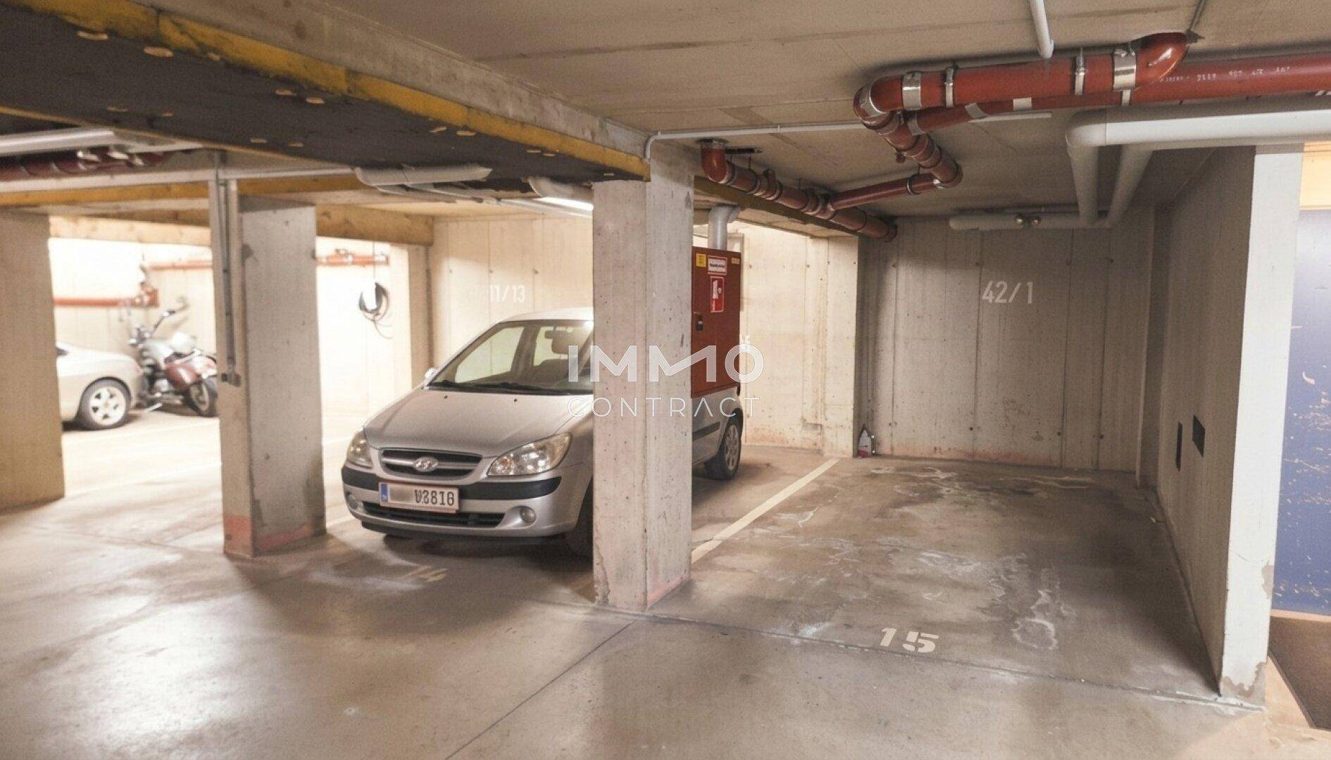 Tiefgarage mit Betonpfeilern, sichtbaren Rohrleitungen und einem geparkten Fahrzeug.