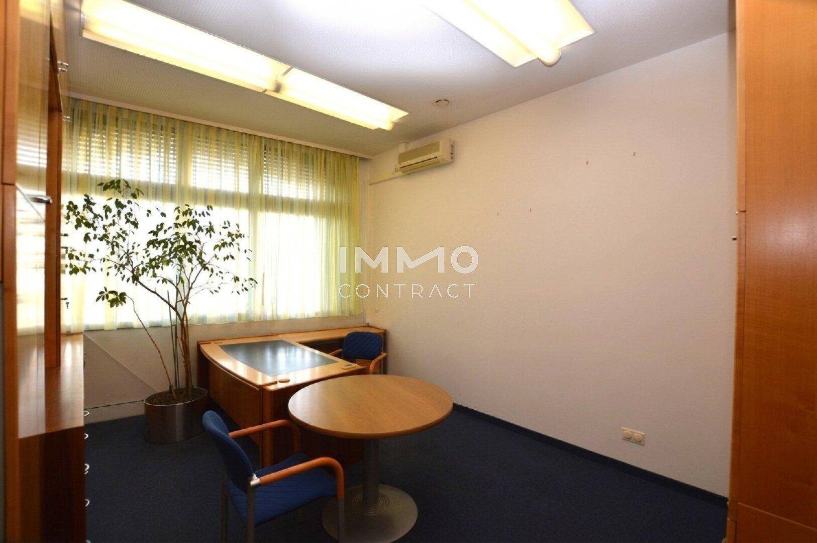 Helles Einzelbüro mit großem Fenster, Schreibtisch, rundem Tisch und Klimaanlage.