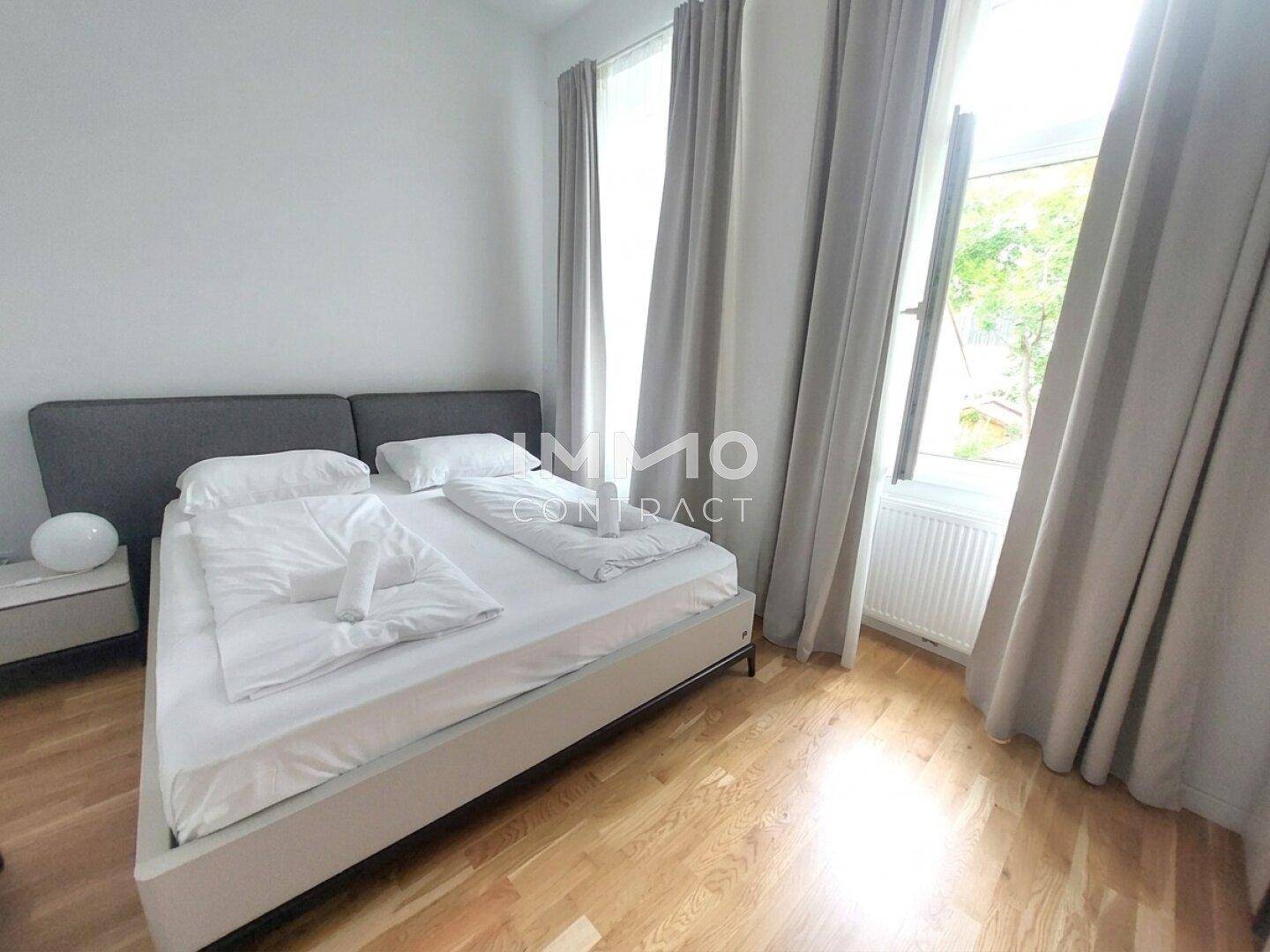 Helles Schlafzimmer mit Doppelbett, Holzboden und großem Fenster, das viel Tageslicht hereinlässt.
