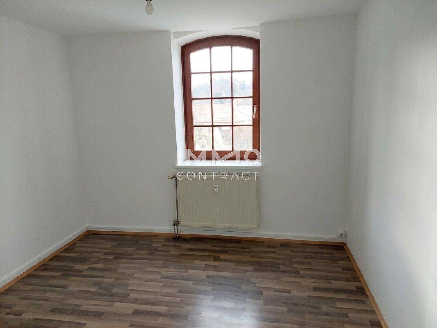 Heller, leerer Raum mit Laminatboden, Heizkörper und einem großen Rundbogenfenster.
