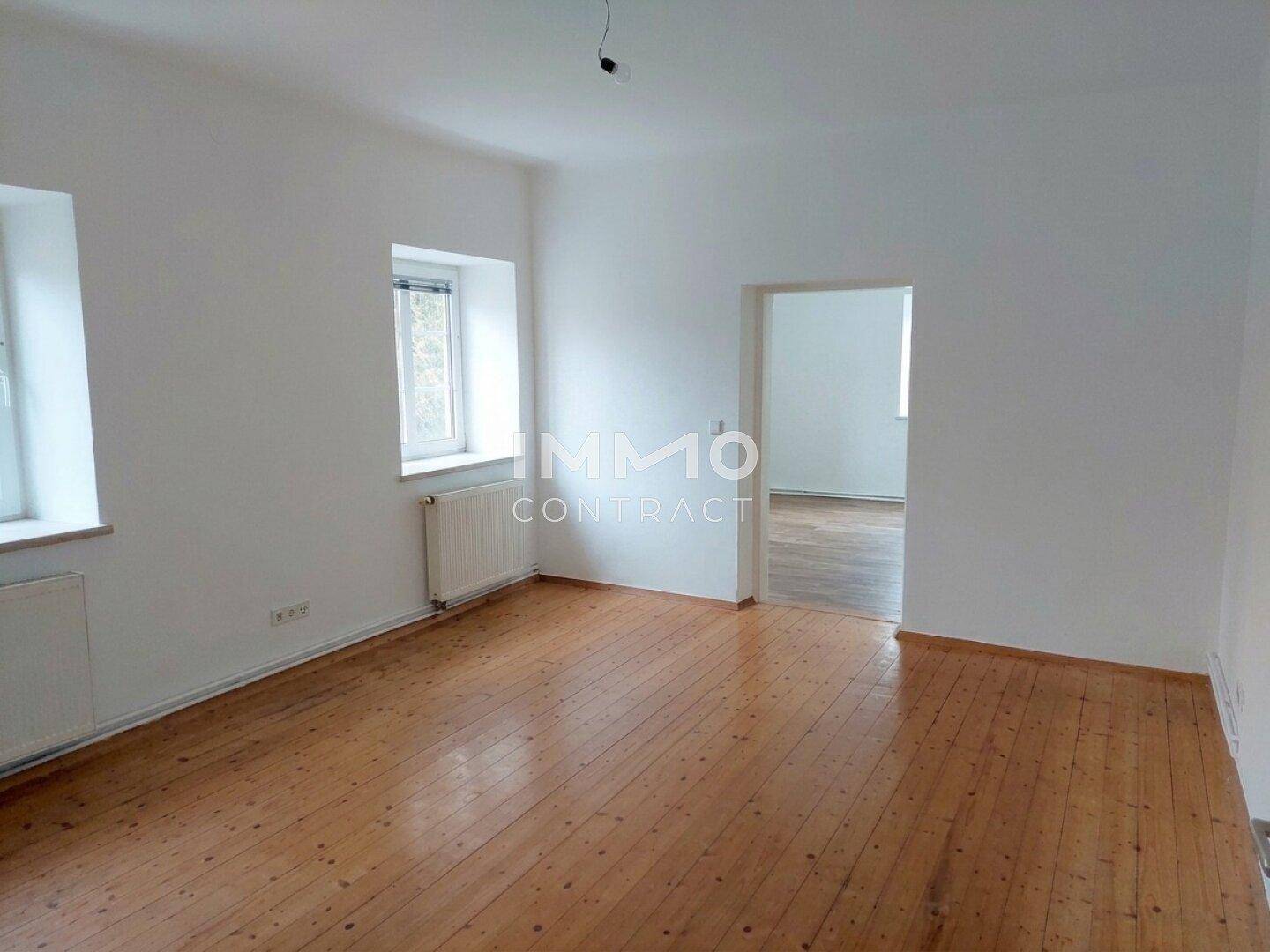 Geräumiges Zimmer mit Holzboden, weißen Wänden und zwei Fenstern, die viel Licht hereinlassen.