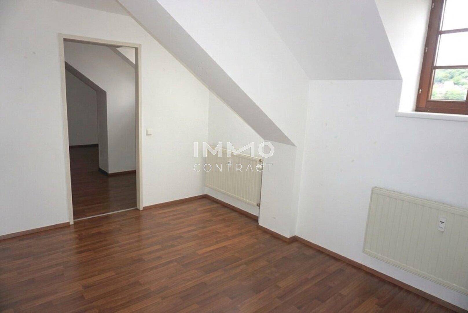Helles Zimmer im Dachgeschoss mit Laminatboden, schrägen Wänden und Zugang zu einem weiteren Raum.