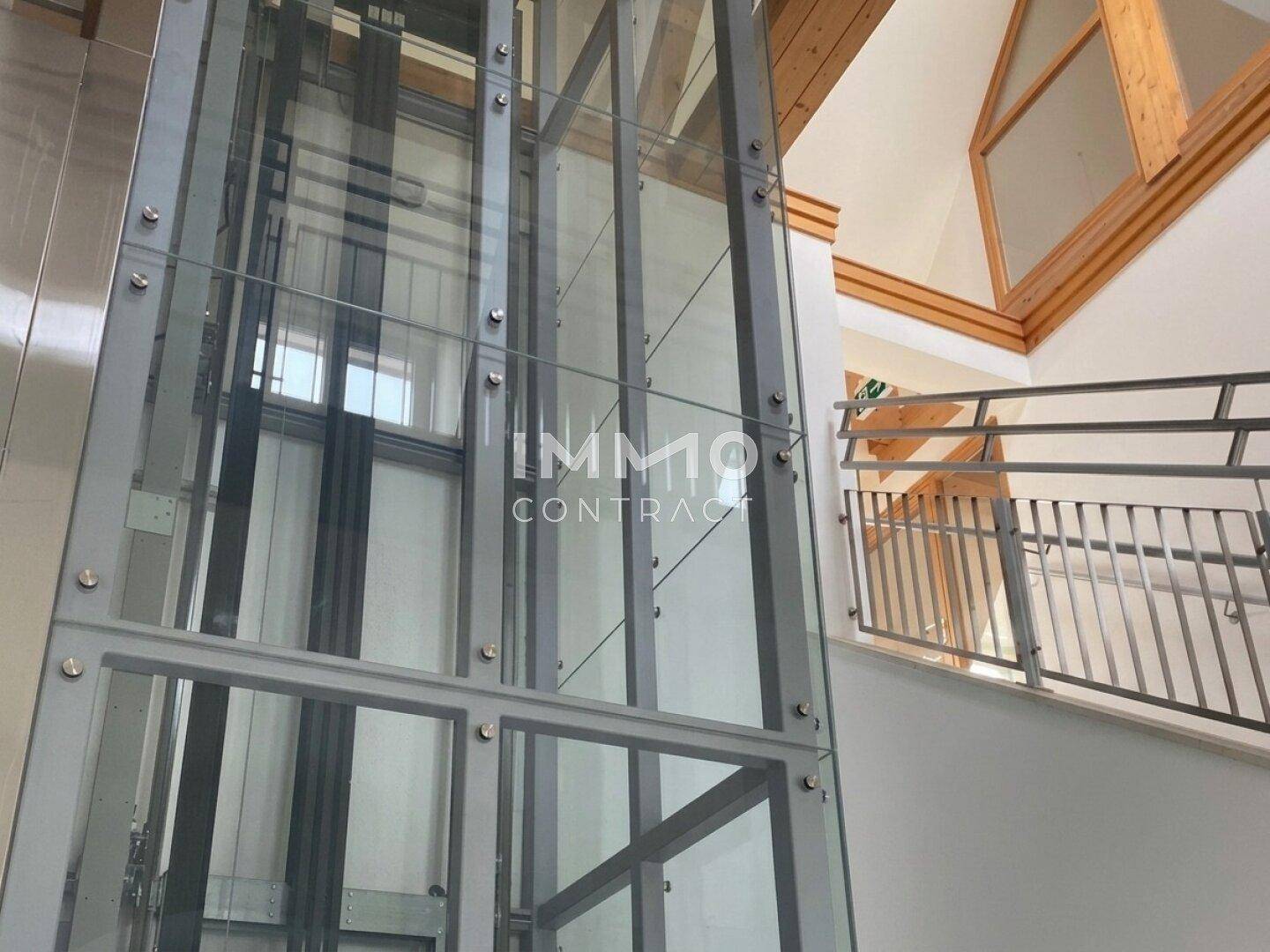 Detailansicht eines modernen Glasaufzugs in einem hellen Treppenhaus mit Holzbalkenstruktur.